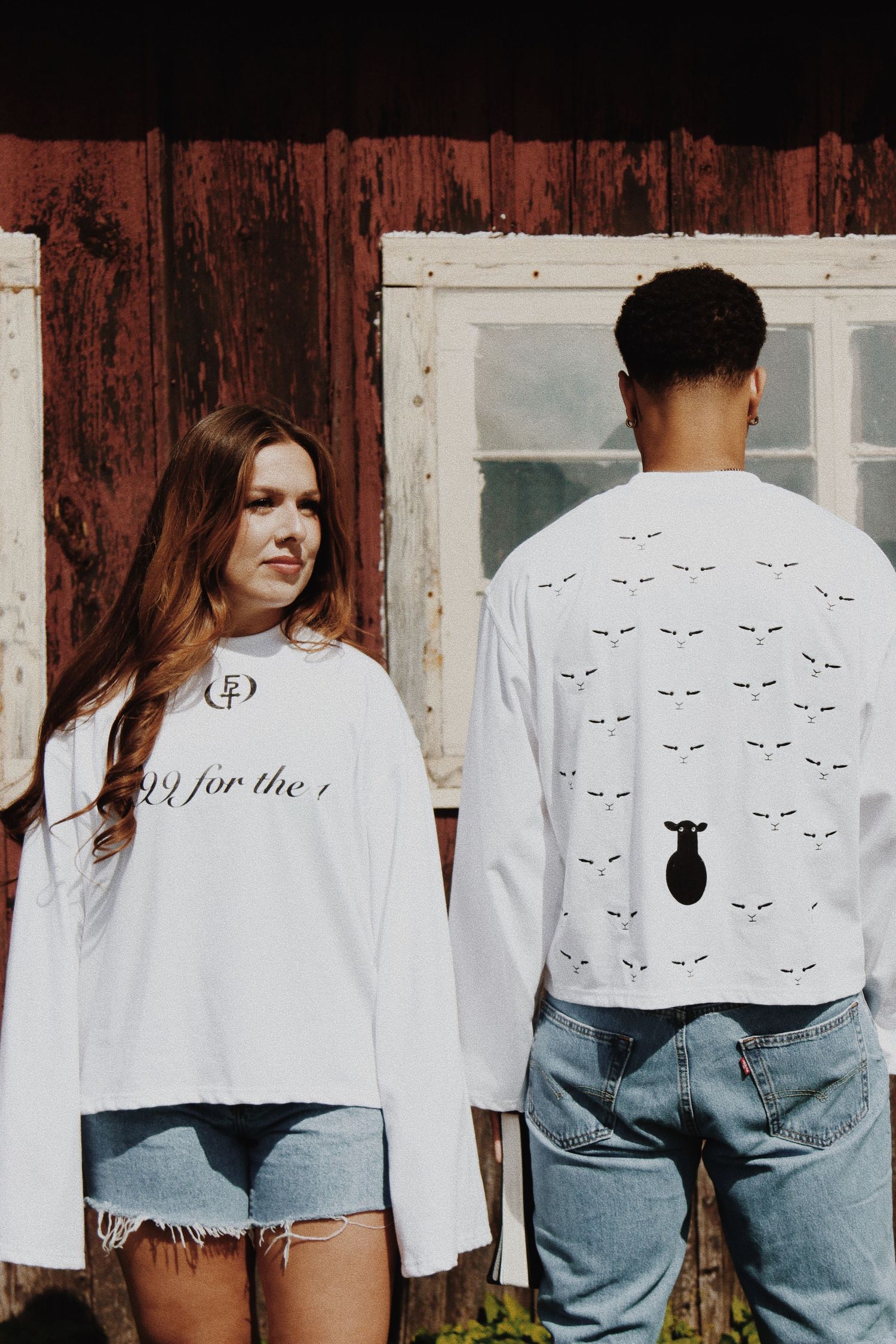 Two models wearing white long-sleeve graphic tees with denim shorts, posing in front of a rustic red barn.
