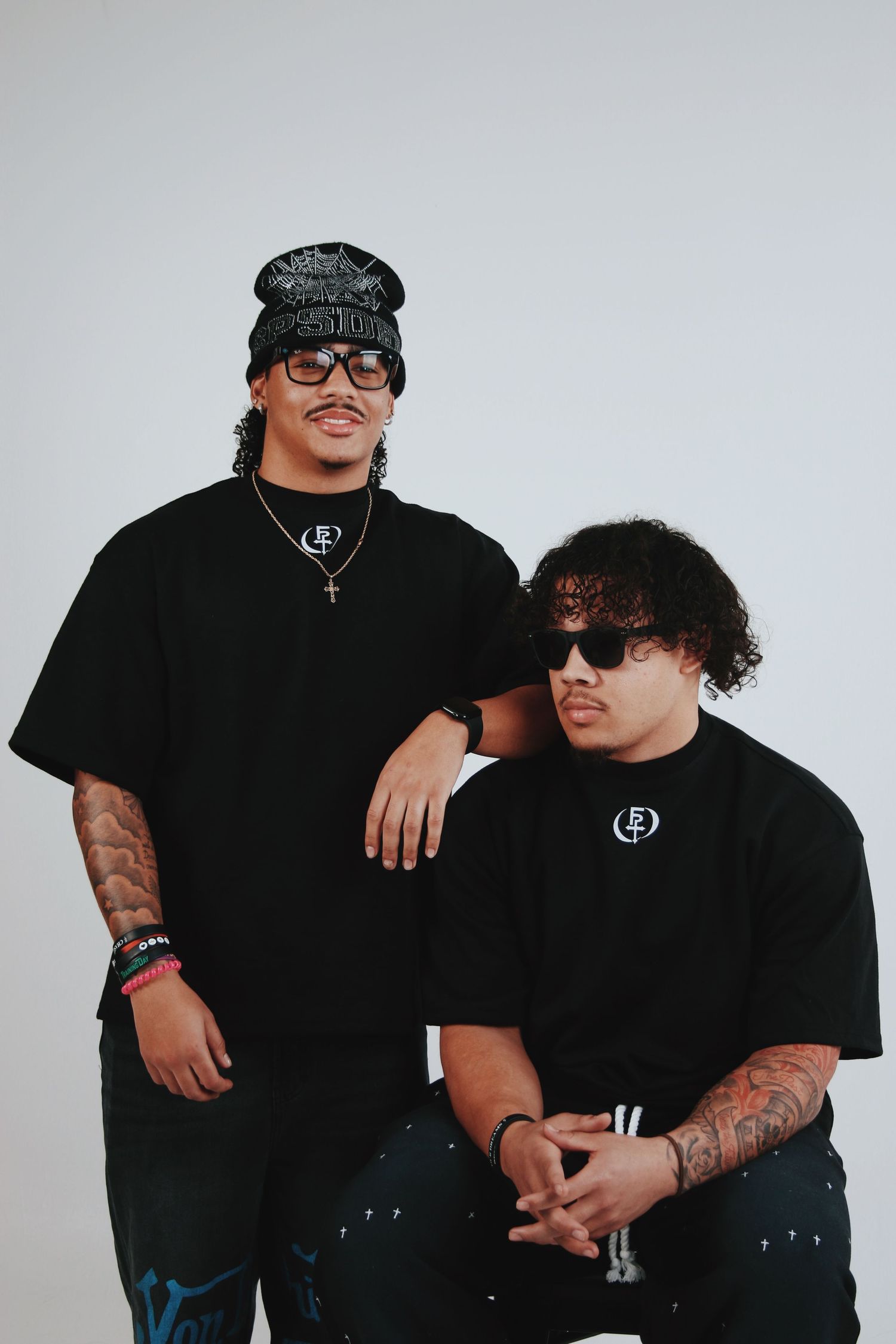 Two young men in matching black graphic tees, sunglasses, and tattoos pose together against a white background.