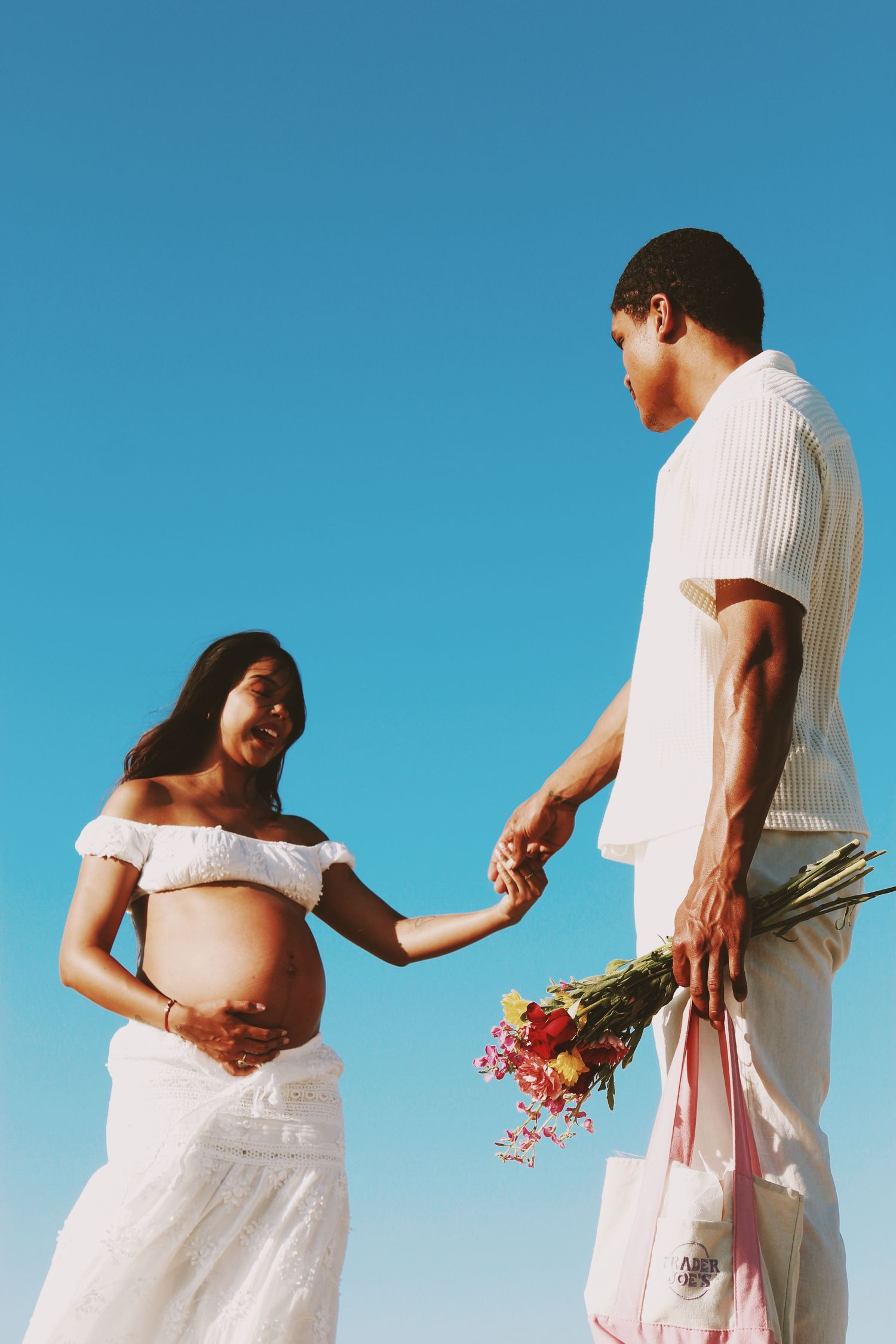 Pregnant woman in white dress holds hands with man offering flowers against clear blue sky.