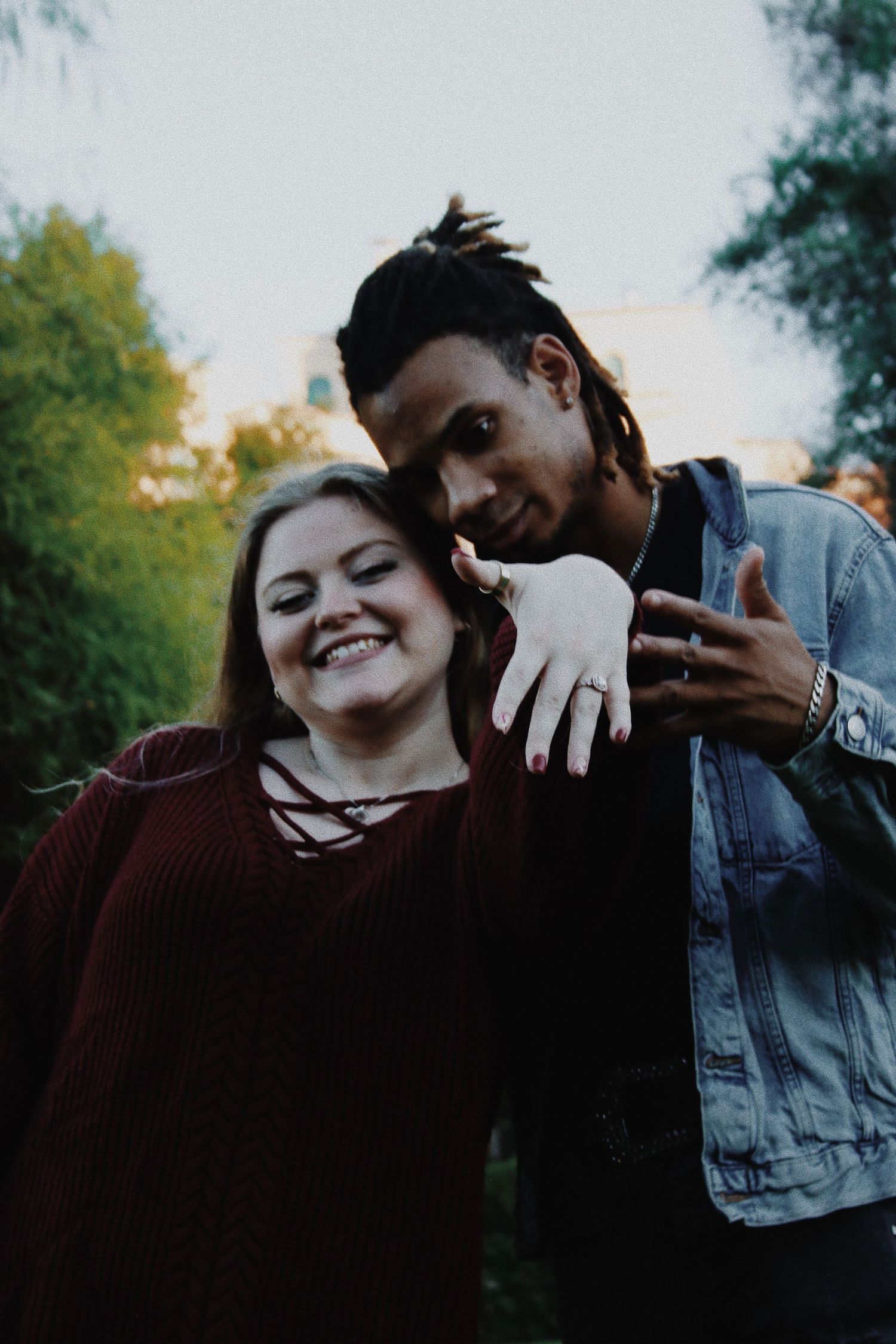 A smiling interracial couple poses outdoors, woman in burgundy sweater leans on mans shoulder as he makes a hand gesture.