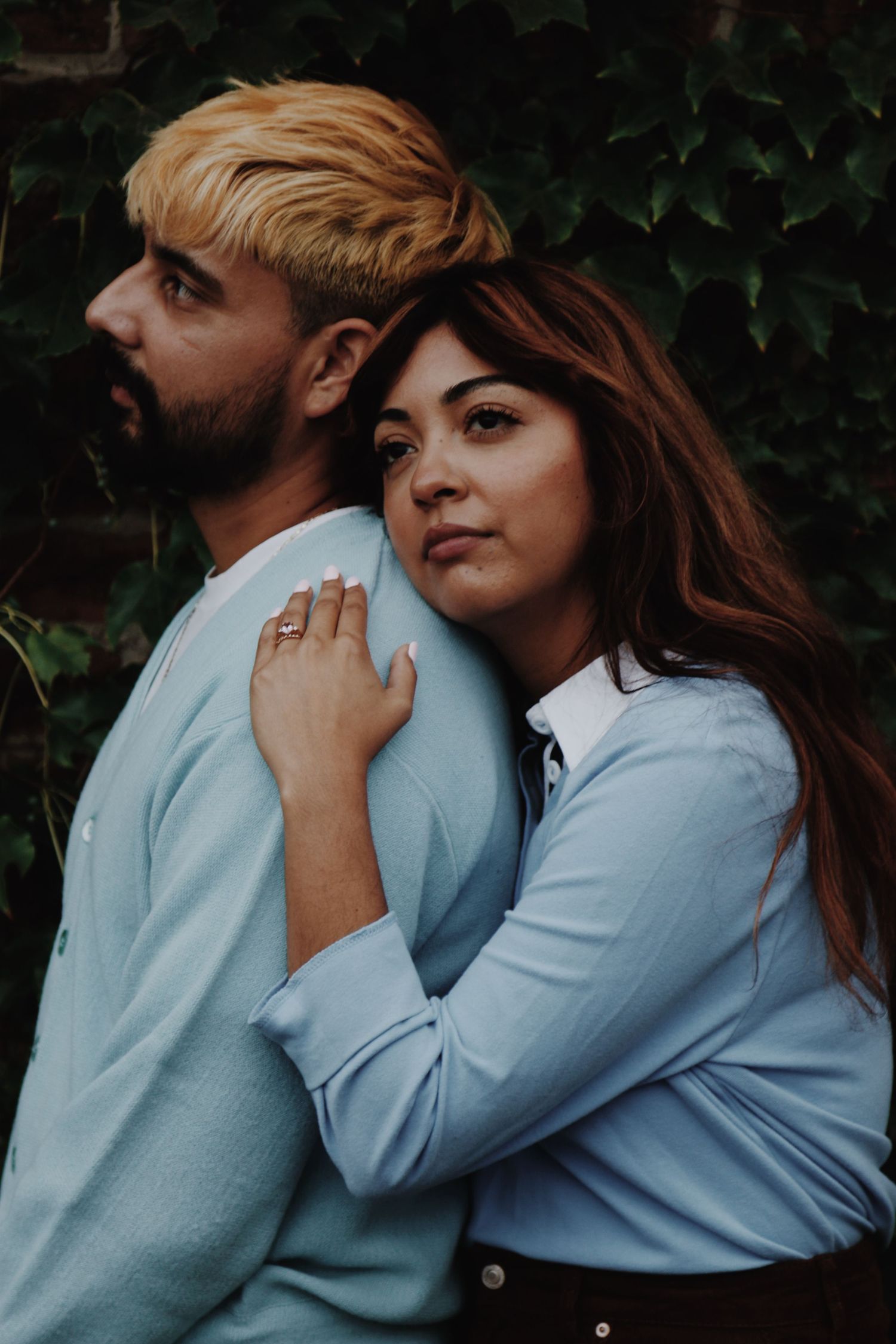 A woman with auburn hair leans on a man with blonde hair, both wearing light blue tops, posed before green ivy.