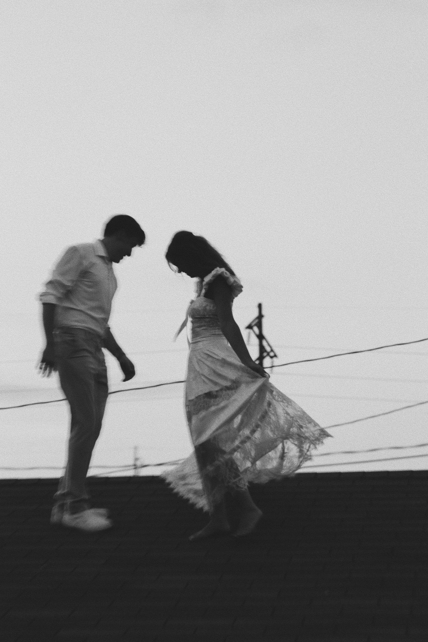 Black and white silhouette of a couple dancing outdoors at dusk, woman in flowing dress, power lines in background.