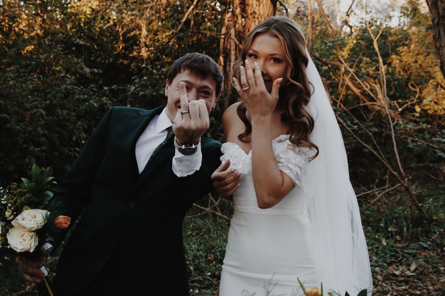 Newlywed couple showing off wedding rings, bride in white gown and groom in dark suit, smiling outdoors.
