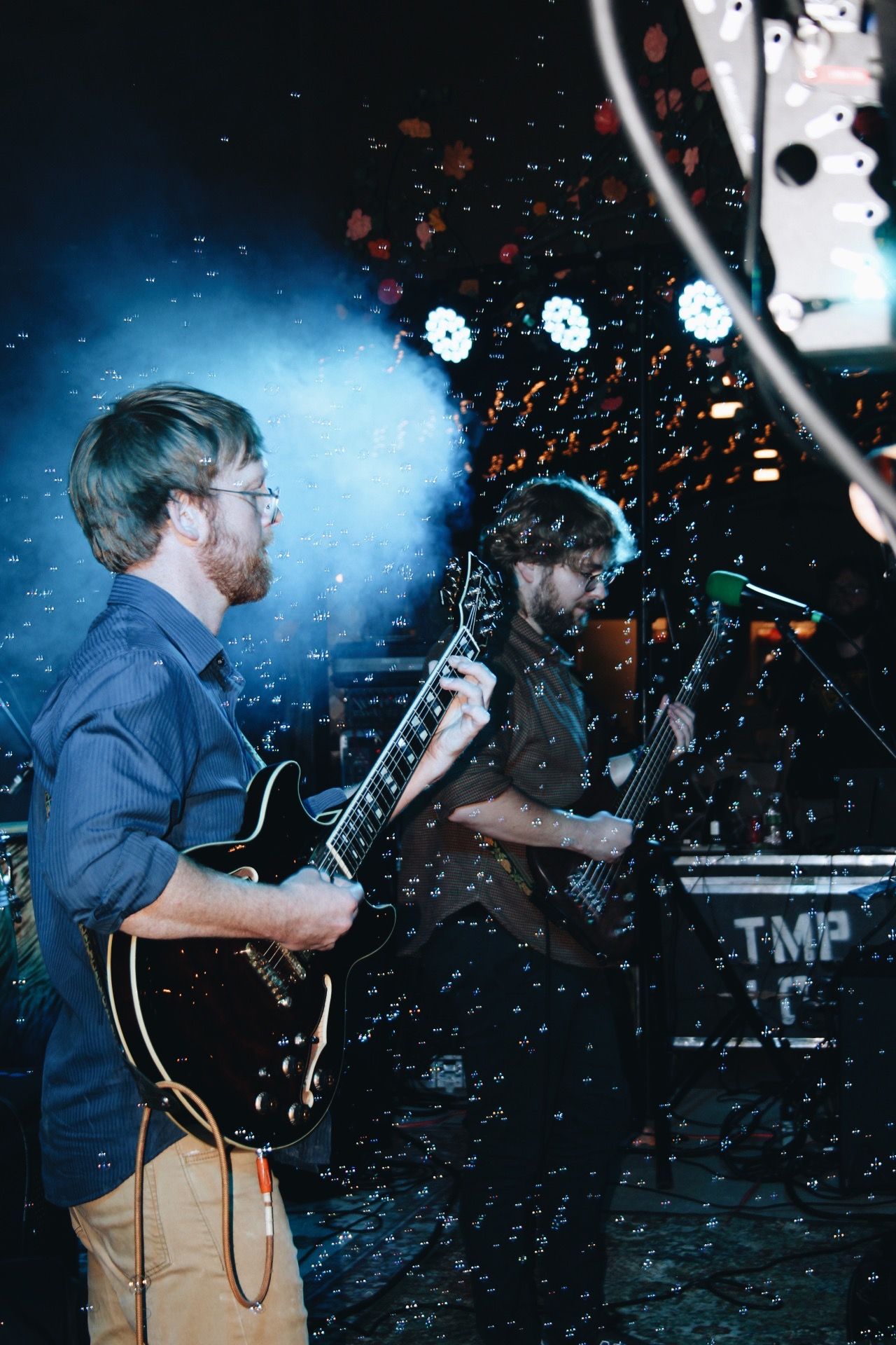 Two male guitarists performing on stage with confetti and blue stage lighting at a live music event.