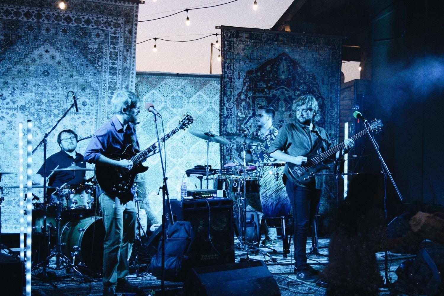 Live band performing on stage with guitars and drums, illuminated by blue stage lighting against decorative tapestry backdrop.