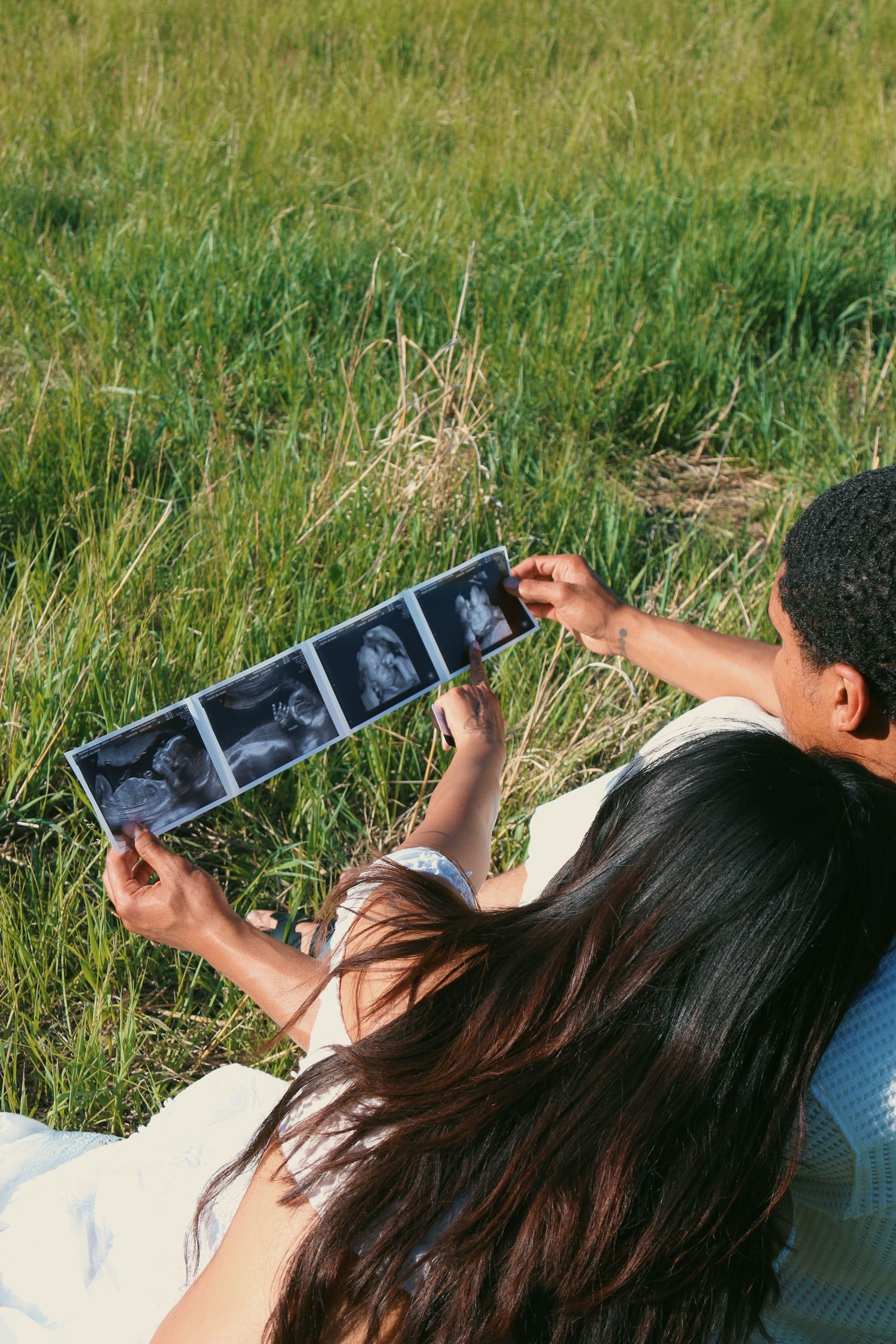 Couple lying in grass holding ultrasound photos outdoors on sunny day.