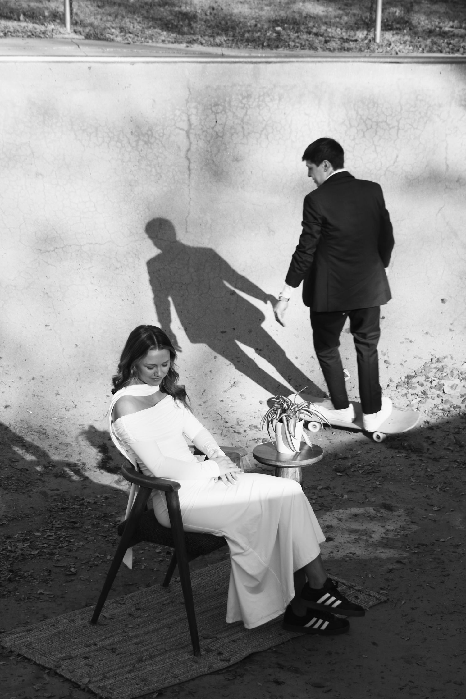 Black and white photo of bride in white dress seated in chair while groom in suit stands near wall with shadows.