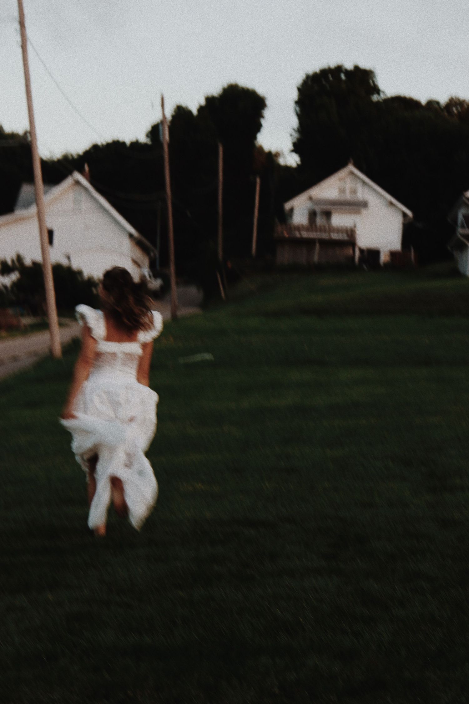 A blurry photo of a young girl in a white dress running across a green lawn near white houses at dusk.