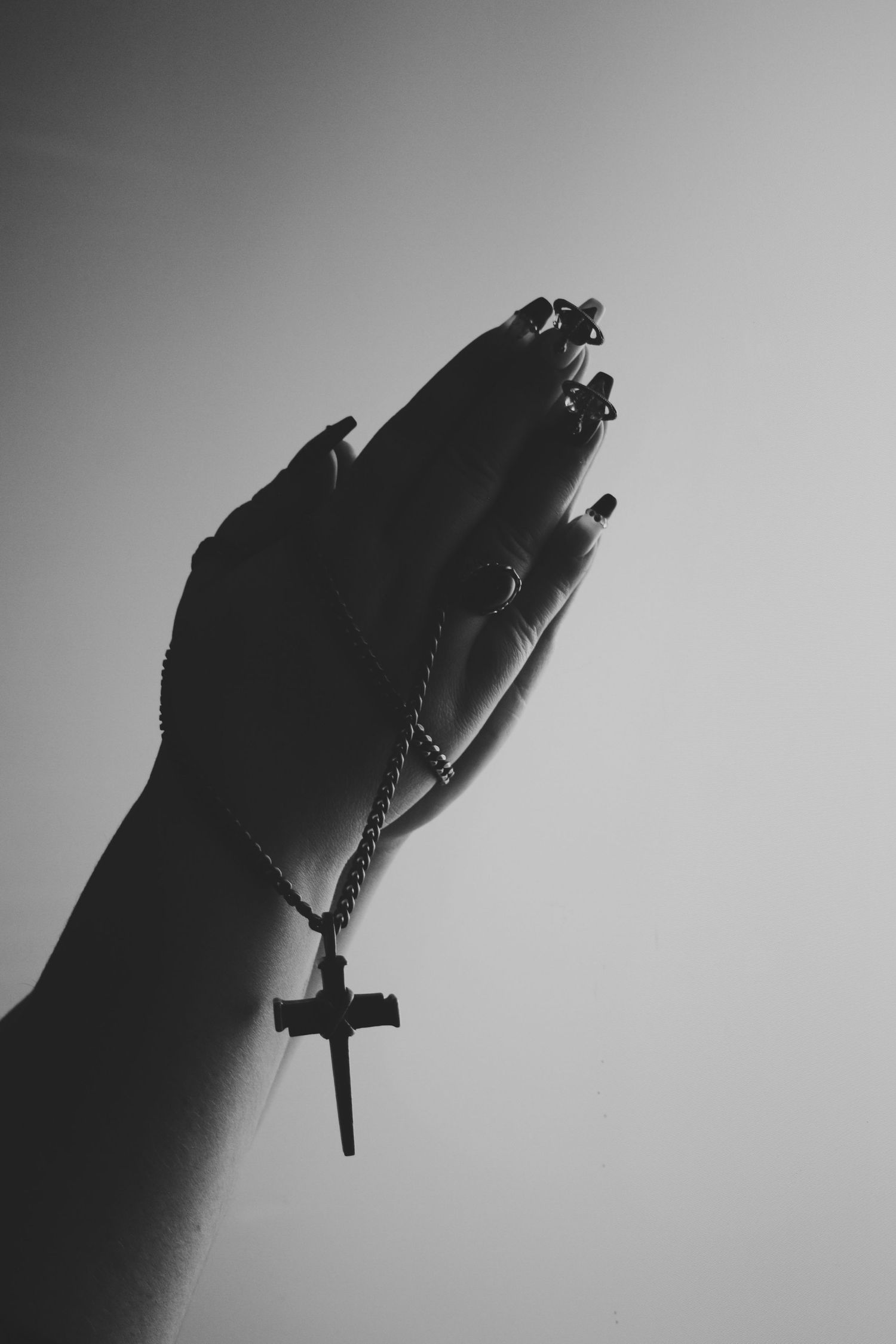 Black and white photo of praying hands holding a rosary cross necklace against a light background.