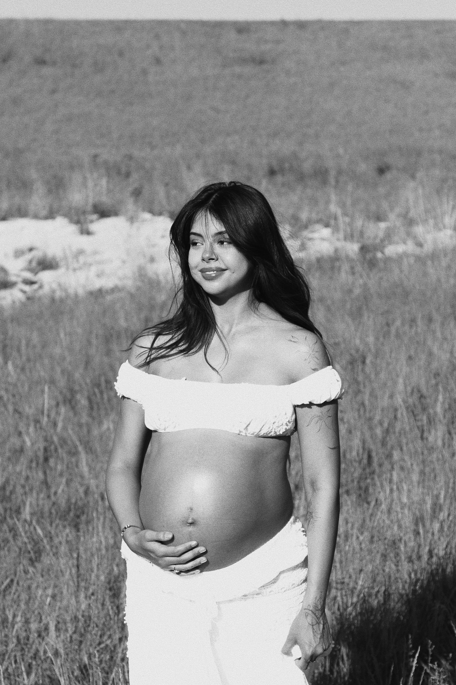 Black and white photo of a smiling pregnant woman in a white crop top holding her baby bump in an outdoor field.