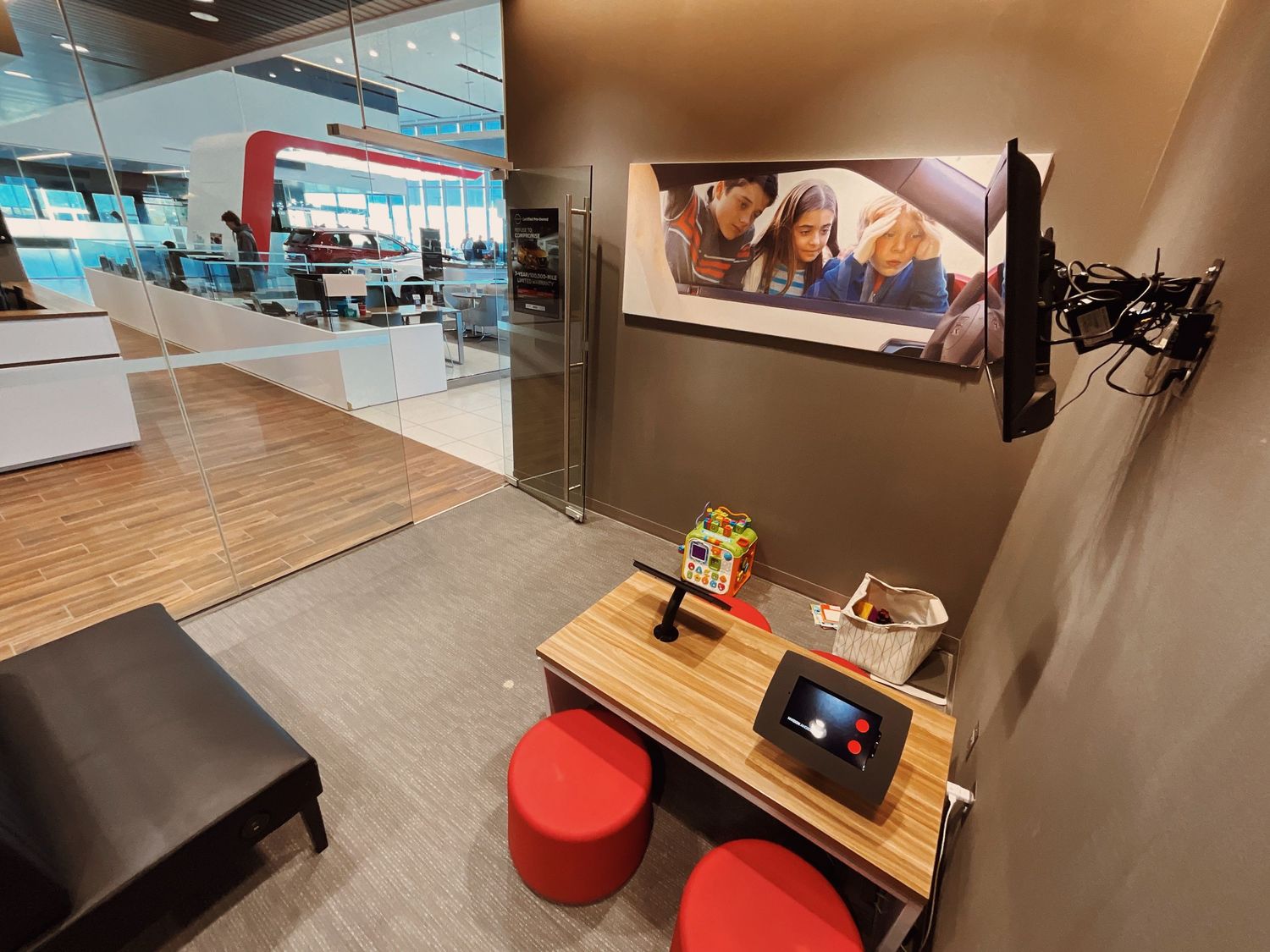 Modern retail waiting area with red stools, wooden table, wall-mounted TV, and colorful family photo in a bright store.