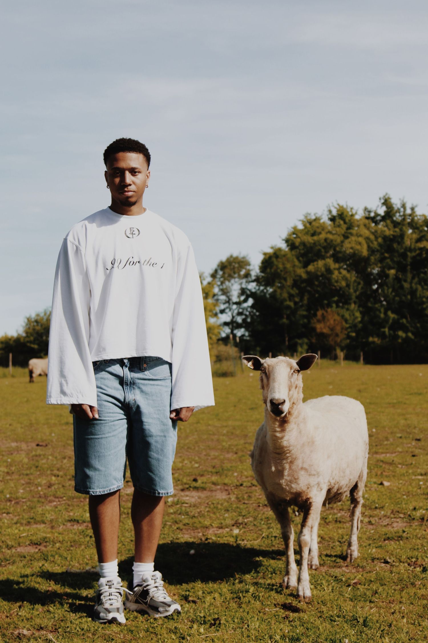 Man in white oversized sweatshirt and denim shorts standing beside a sheep on a sunny farm.