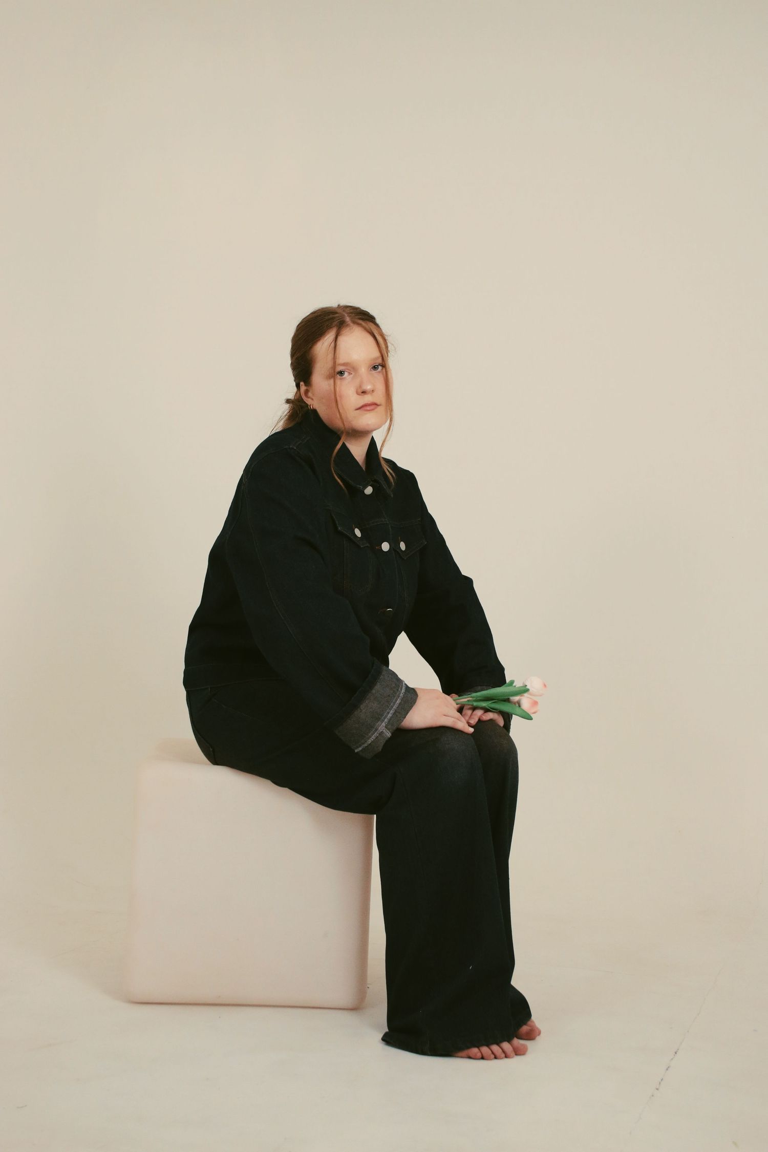 Woman in black denim outfit seated on white cube holding green stems, barefoot against neutral background.