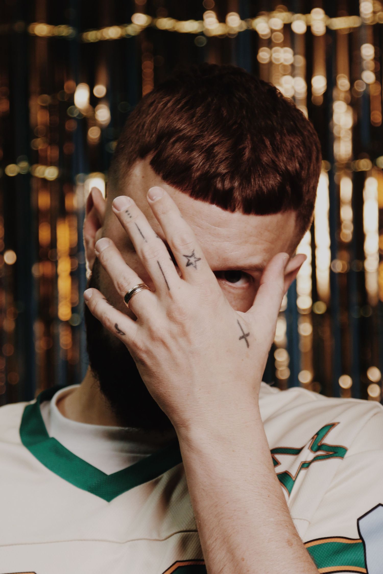 Young man with finger tattoos and a ring covers his face with his hand, wearing a white jersey with bokeh lights behind.