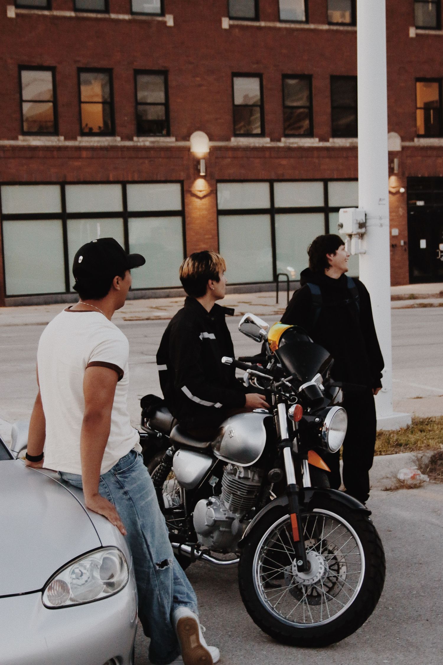 Three young men gathered around a vintage silver motorcycle on an urban street with a red brick building backdrop.