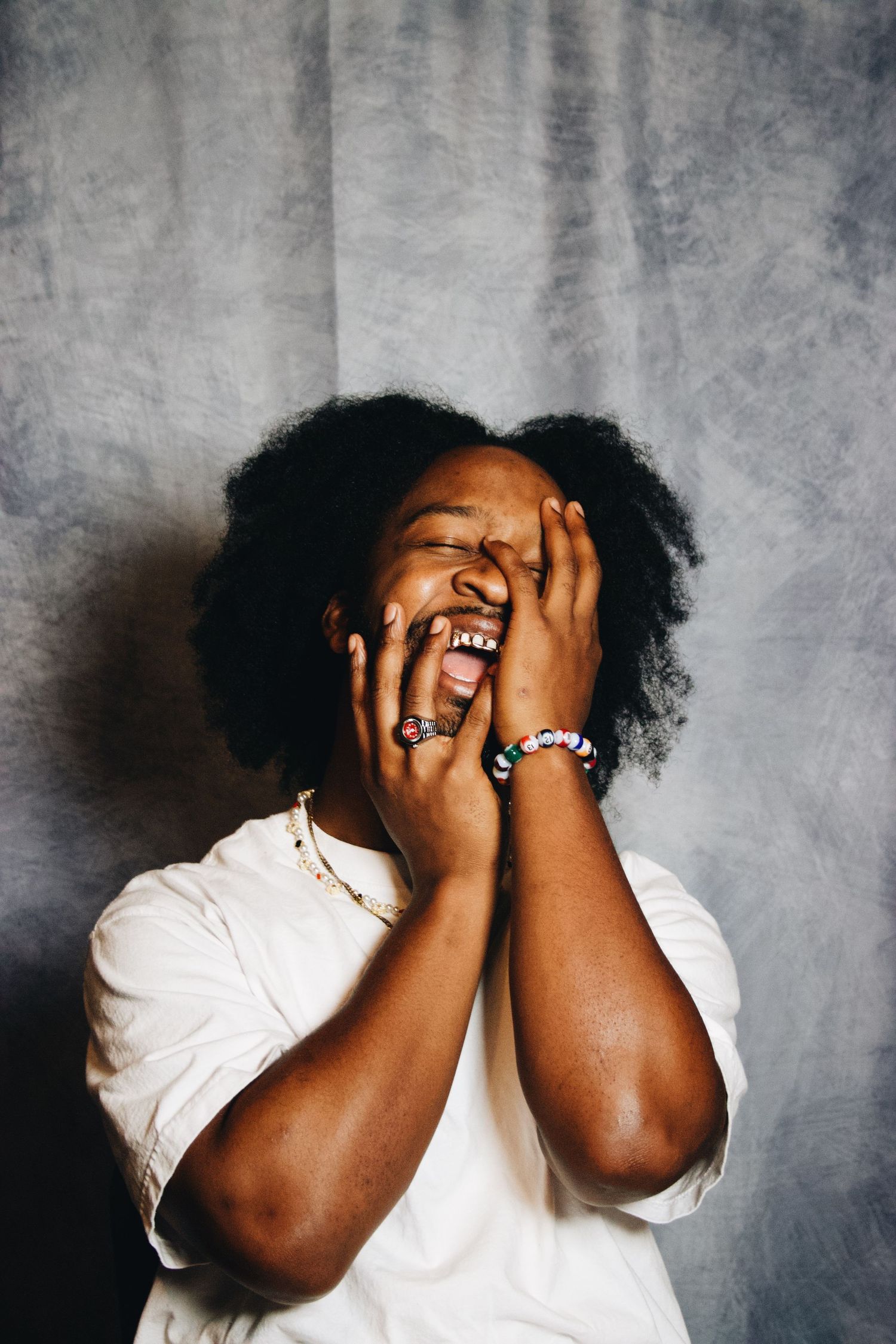 A joyful person with natural hair laughs with hands covering face, wearing bracelets and white tee against gray backdrop.
