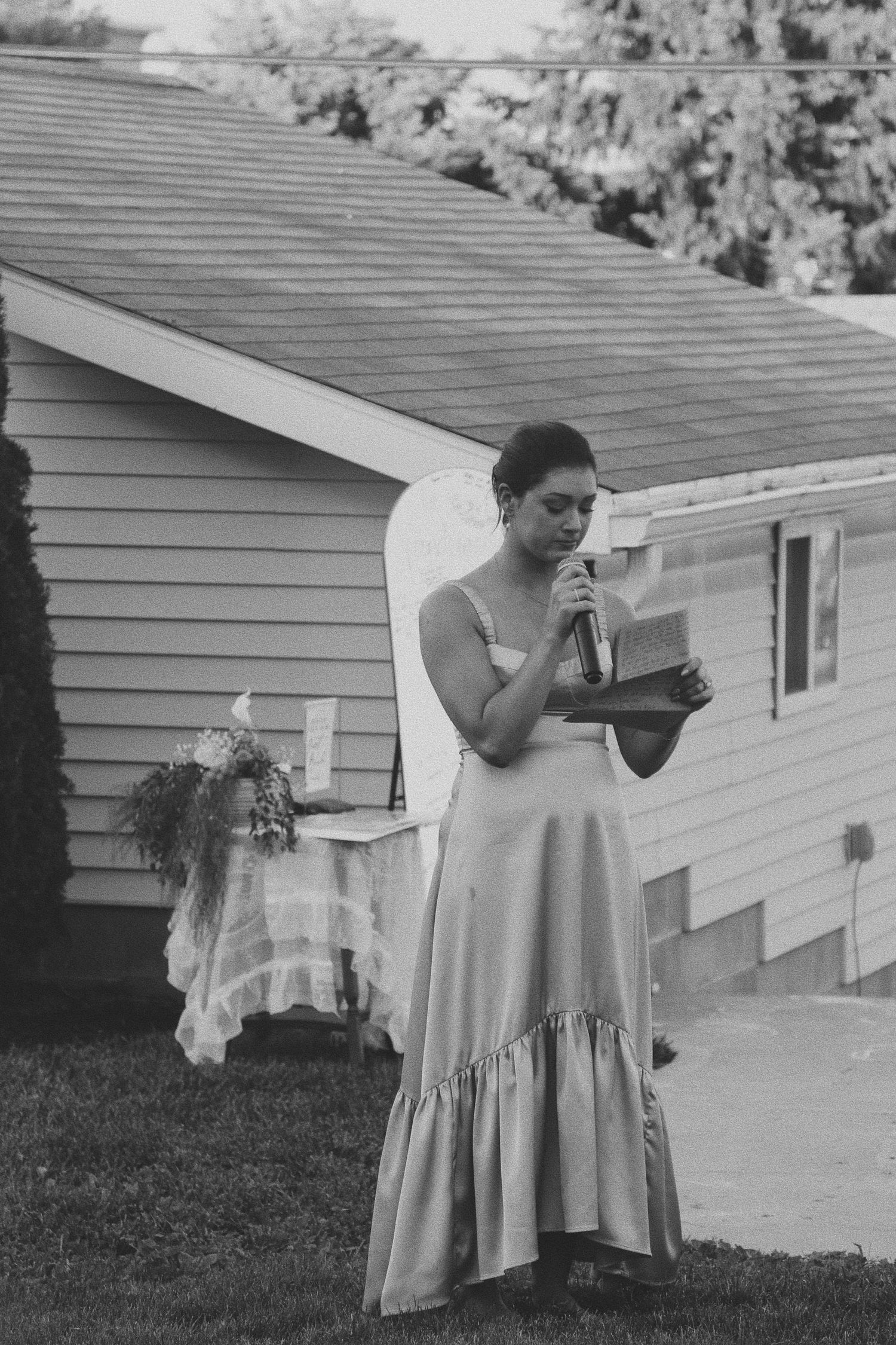 Woman in elegant gown reads from paper at outdoor wedding ceremony, black and white photo.