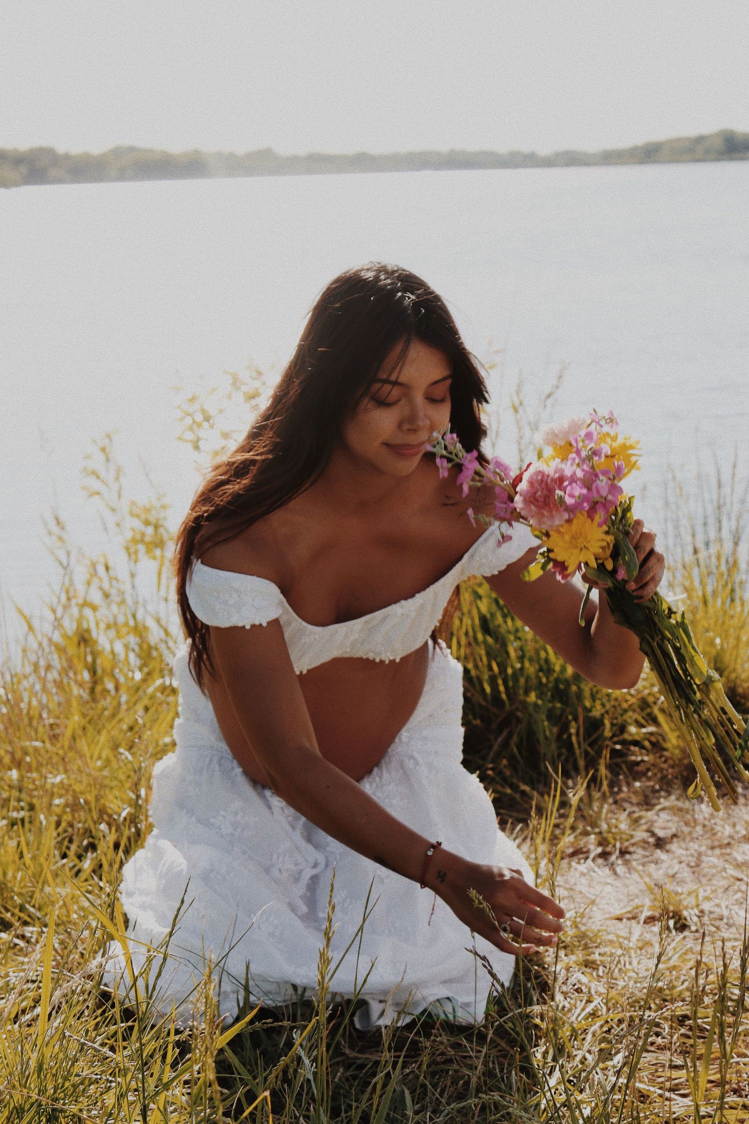 A brunette woman in a white off-shoulder dress smells a bouquet of wildflowers while sitting by a lake.