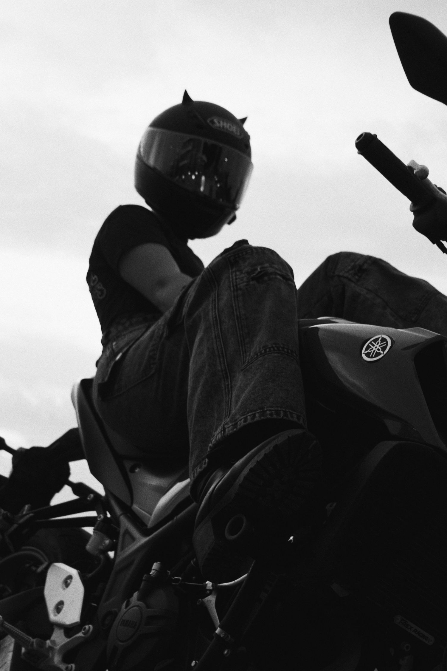 Black and white photo of a motorcyclist in a helmet sitting on a Yamaha motorcycle, shot from a low angle.