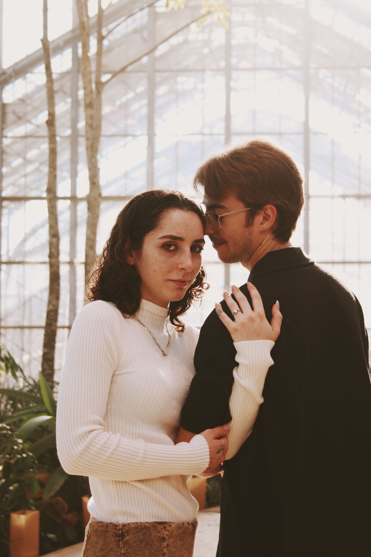 Couple embracing tenderly in a sunlit greenhouse, woman in white turtleneck sweater and man in black jacket.