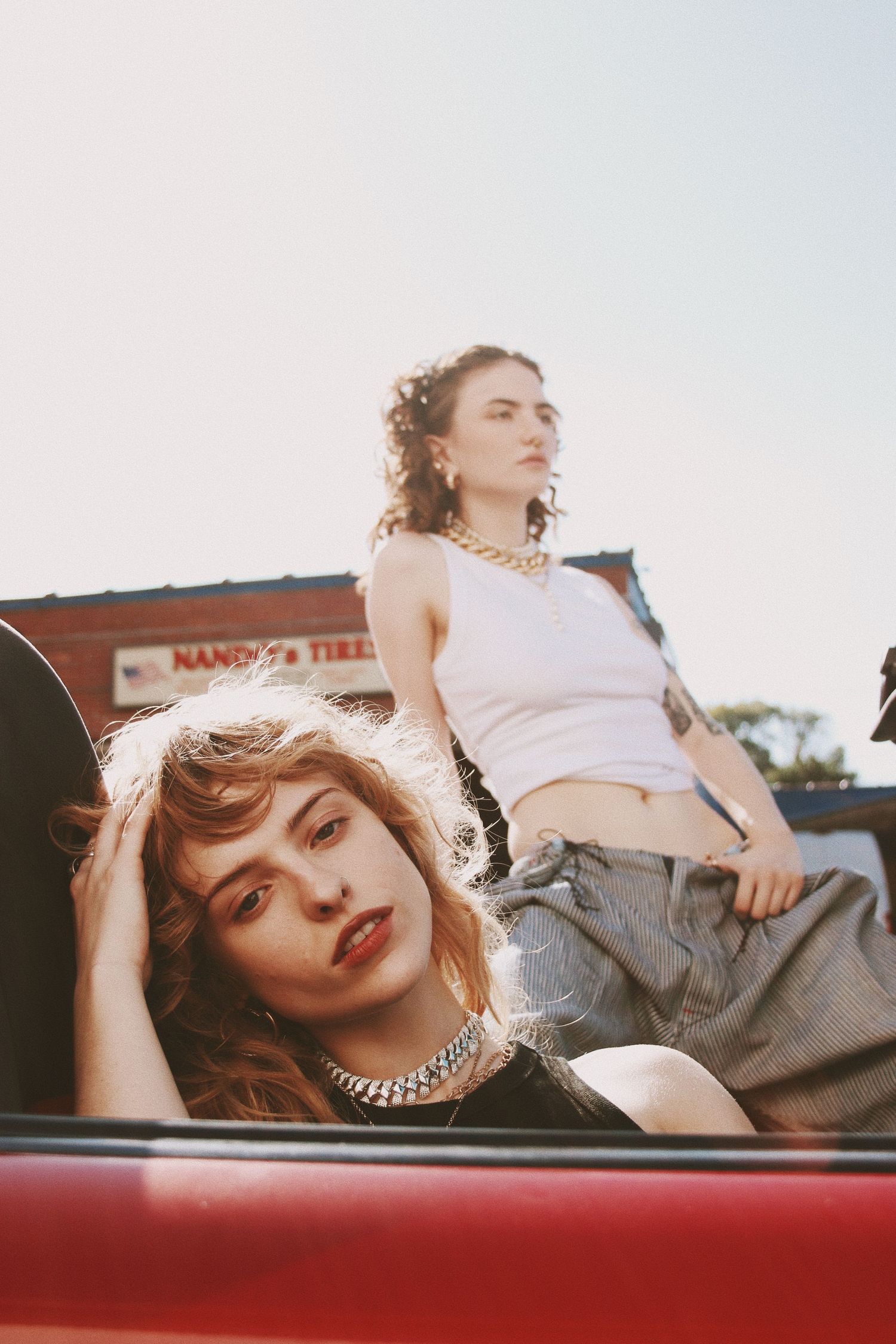 Two young women posing in a red convertible car on a sunny day, wearing casual streetwear and chain necklaces.