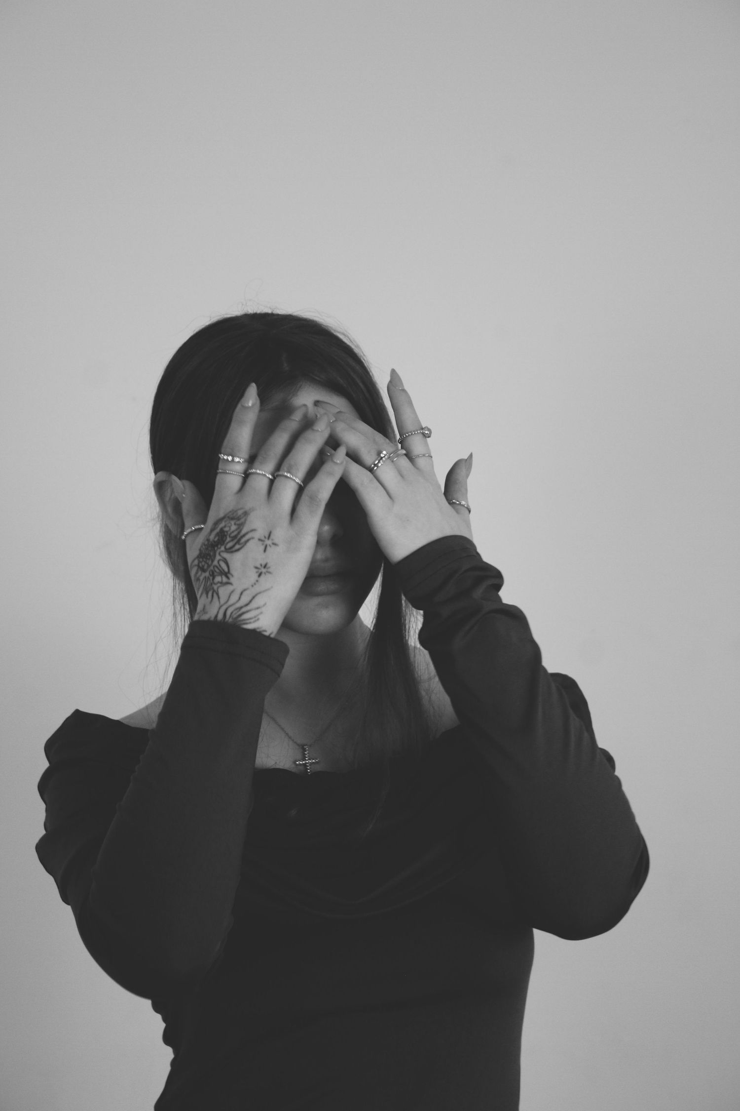 Black and white photo of a woman covering her face with tattooed hands adorned with rings, wearing a dark top.