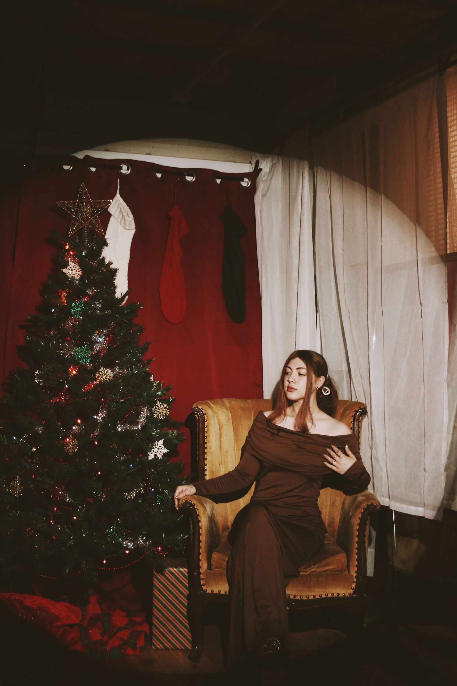 Woman in brown dress sits in velvet chair beside decorated Christmas tree with stockings and red curtains.