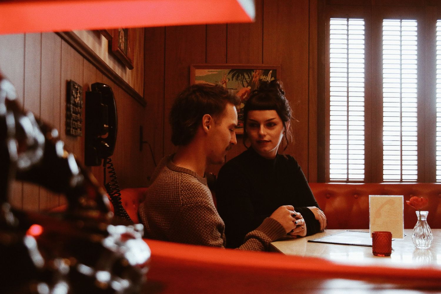 Couple sitting together in a cozy retro diner booth, engaged in intimate conversation near a vintage red telephone.