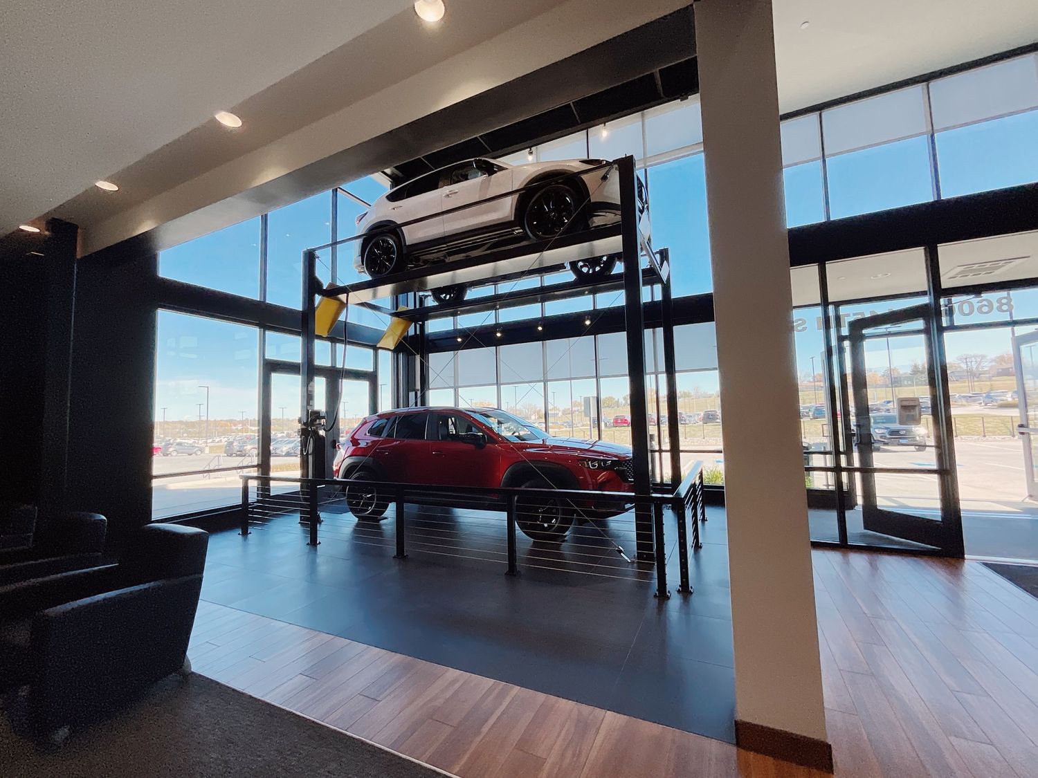 Car dealership showroom with a two-level car lift displaying vehicles, featuring modern wood floors and large windows.