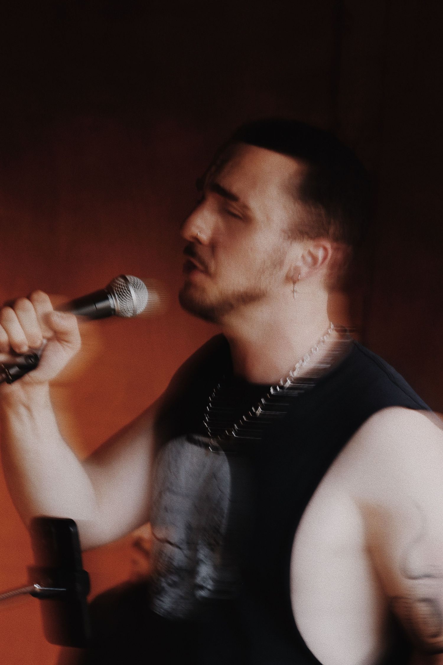 A male rock singer performing live, holding a microphone, wearing a black sleeveless shirt and chain necklace.