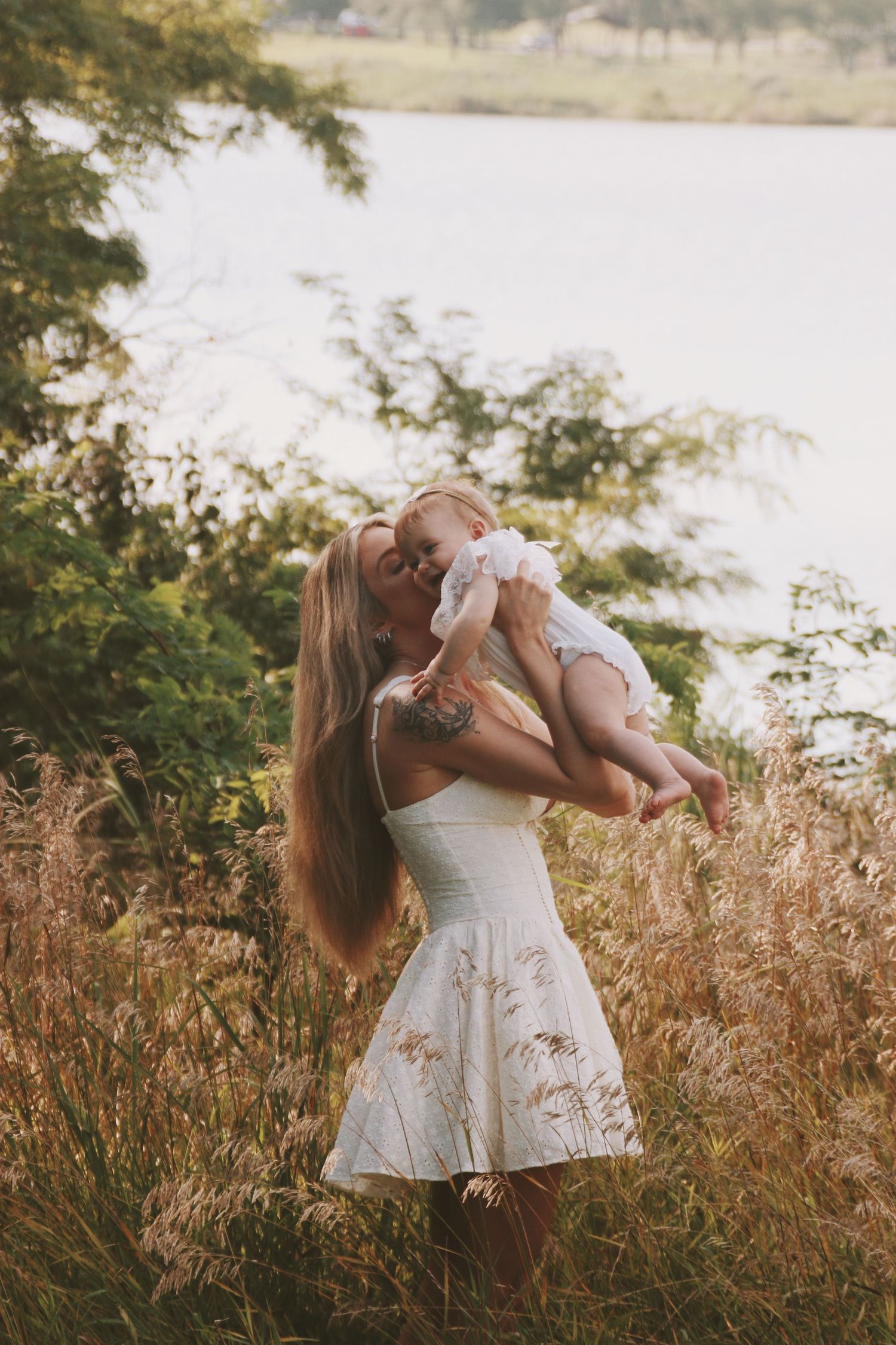 Woman in white dress kissing baby outdoors near lake surrounded by tall golden grass and green trees.