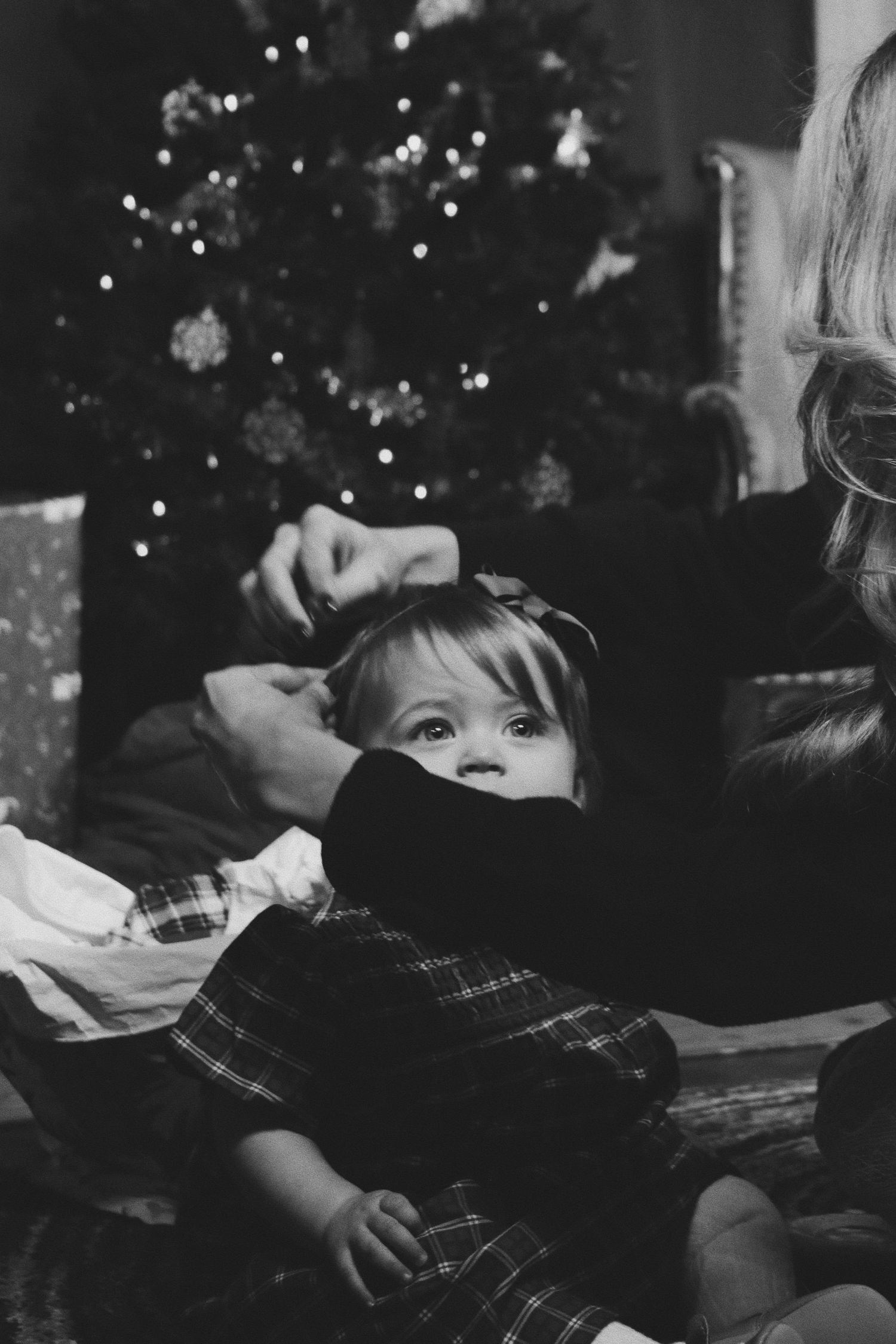 Black and white photo of a toddler girl in plaid dress near a Christmas tree with bokeh lights.