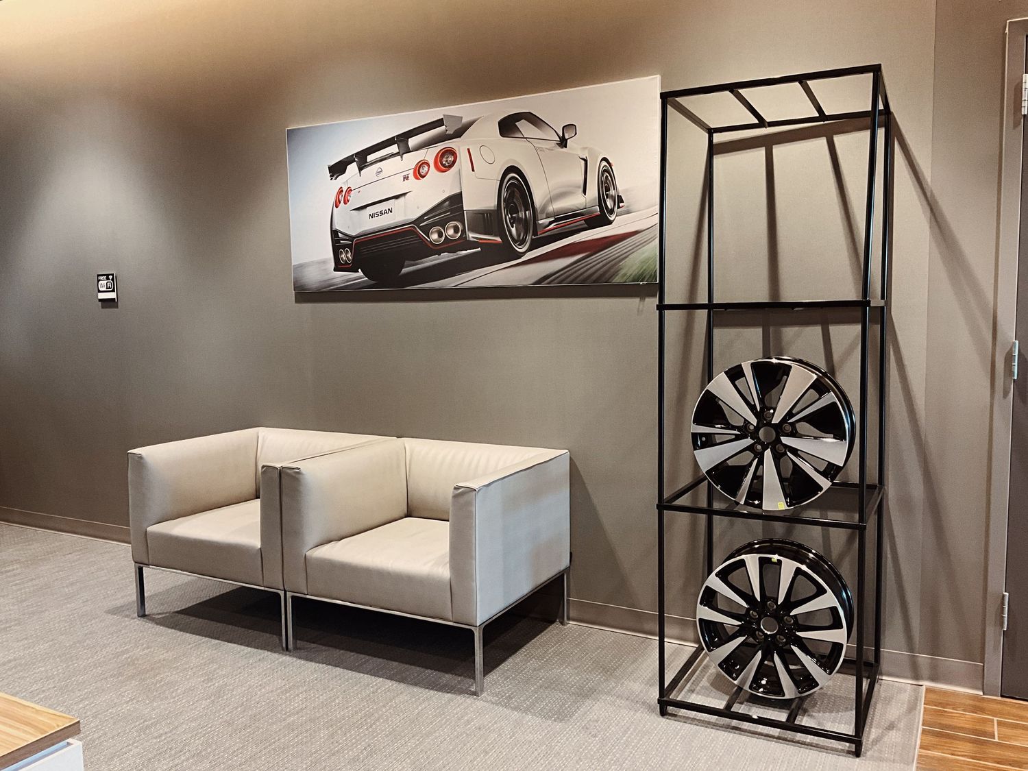Nissan GT-R wall art above two white chairs beside a metal shelf displaying stylish alloy wheels in a modern showroom.