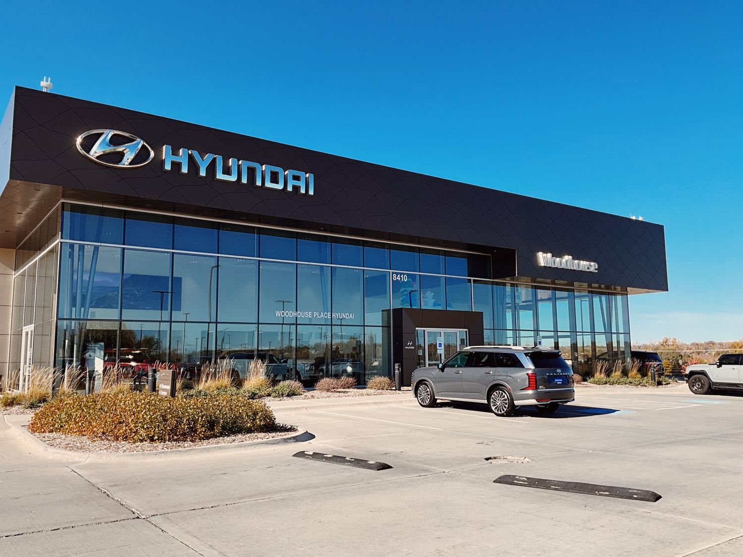 Modern Hyundai car dealership exterior with large glass facade, black signage, and vehicles parked in the lot under a clear blue sky.