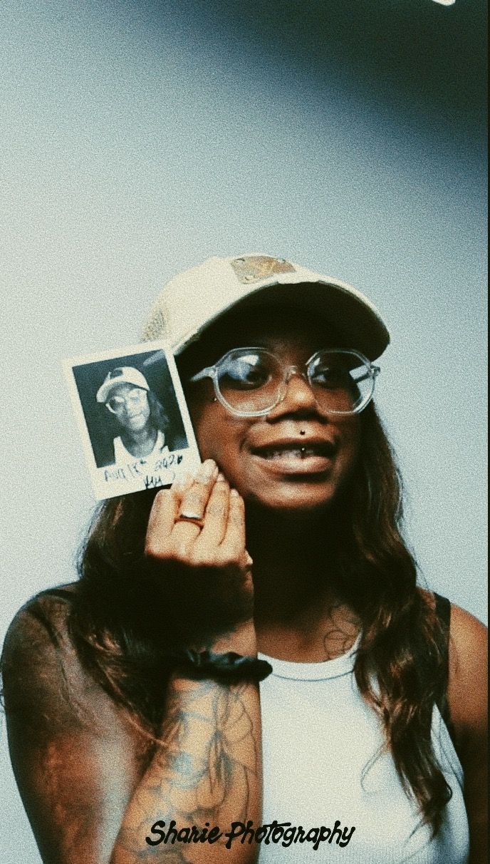 A tattooed woman wearing glasses and a cap holds up a Polaroid photo, smiling, with a nose piercing visible.