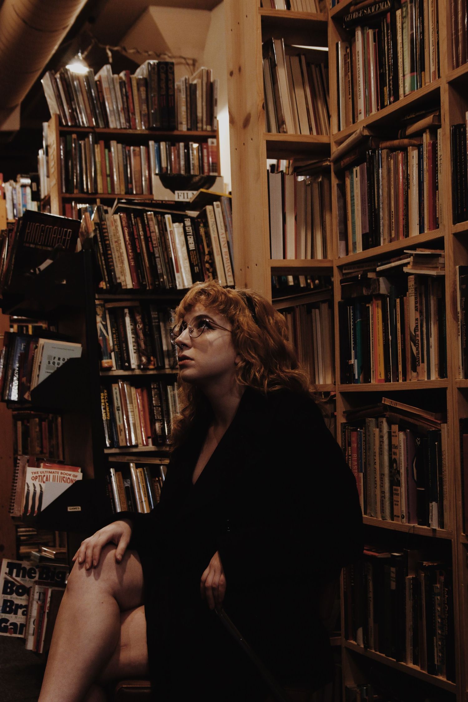 Woman with glasses sitting among tall wooden bookshelves filled with books in a cozy bookstore.