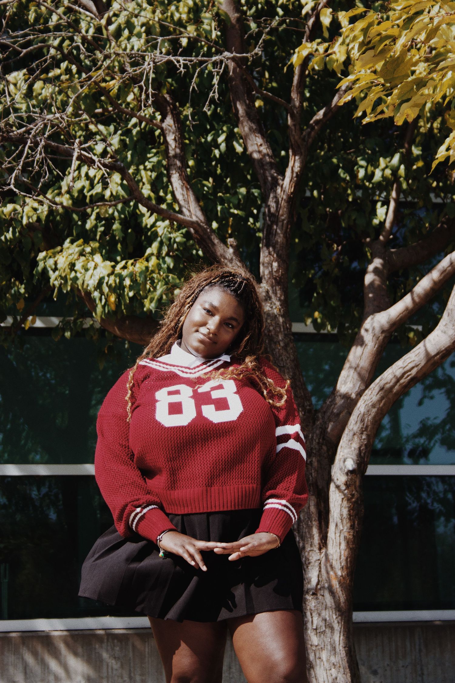 Young Black woman in red #83 varsity sweater and black skirt poses outdoors near a tree in sunlight.