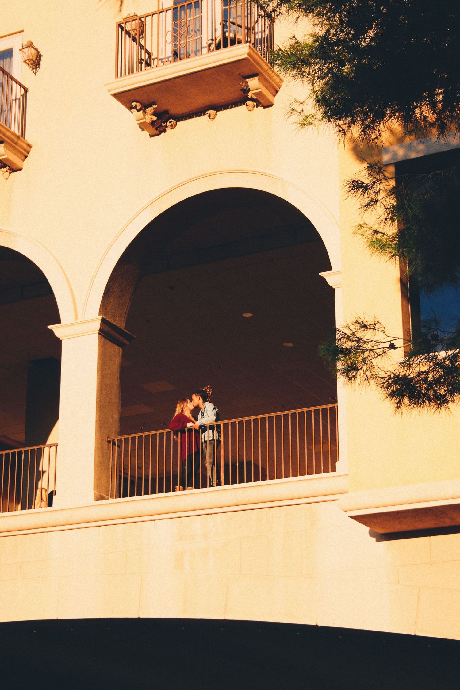 Couple sharing a romantic moment on an arched balcony of a golden Mediterranean-style building at sunset.