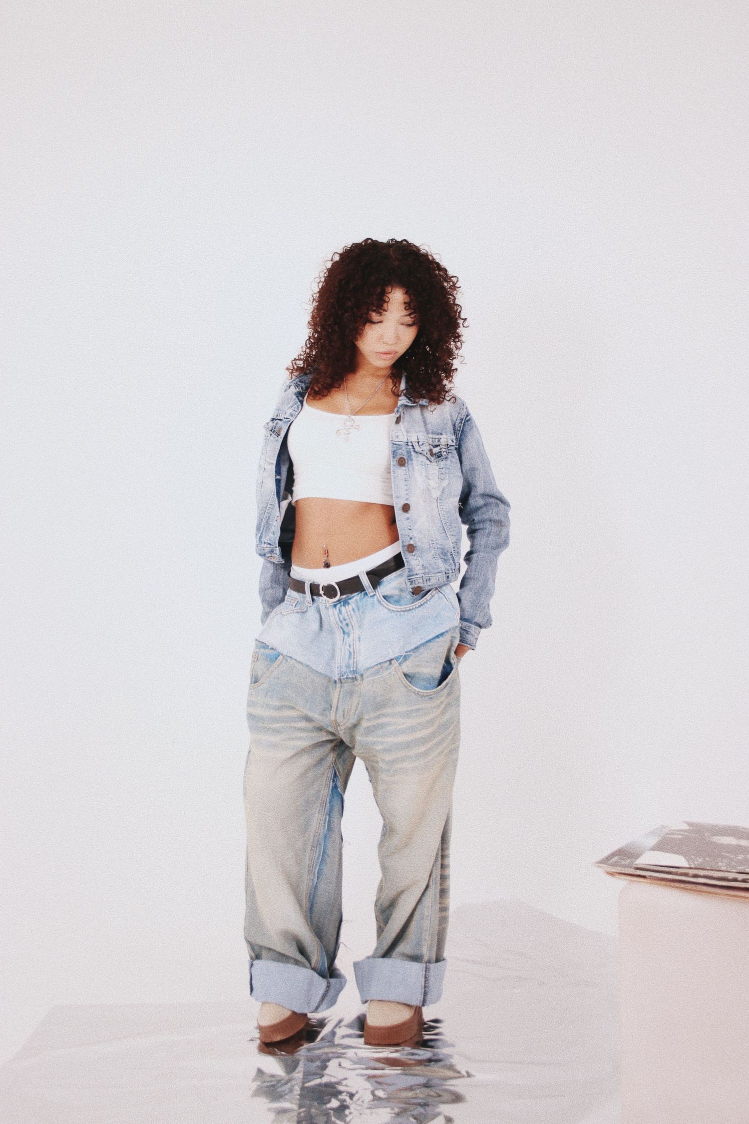 Young woman with curly hair wearing a white crop top, denim jacket, and oversized light-wash jeans with a black belt.