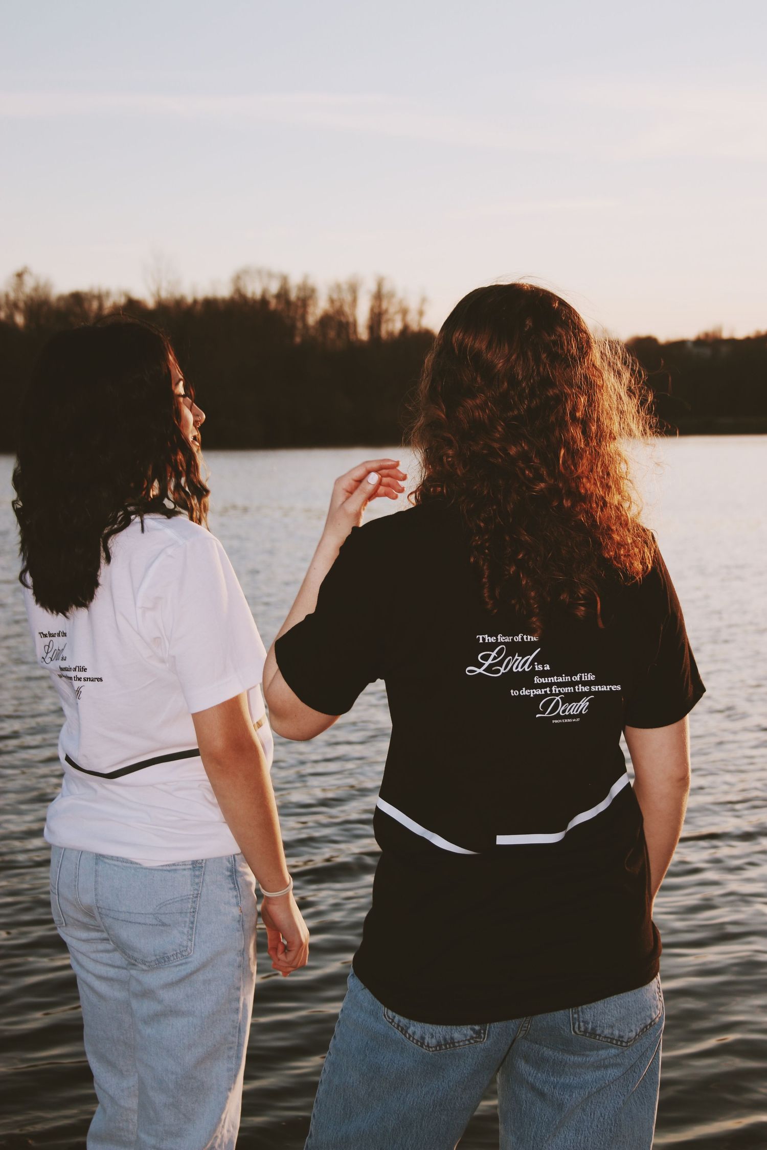 Two women standing by a lake at sunset, wearing graphic t-shirts, viewed from behind.