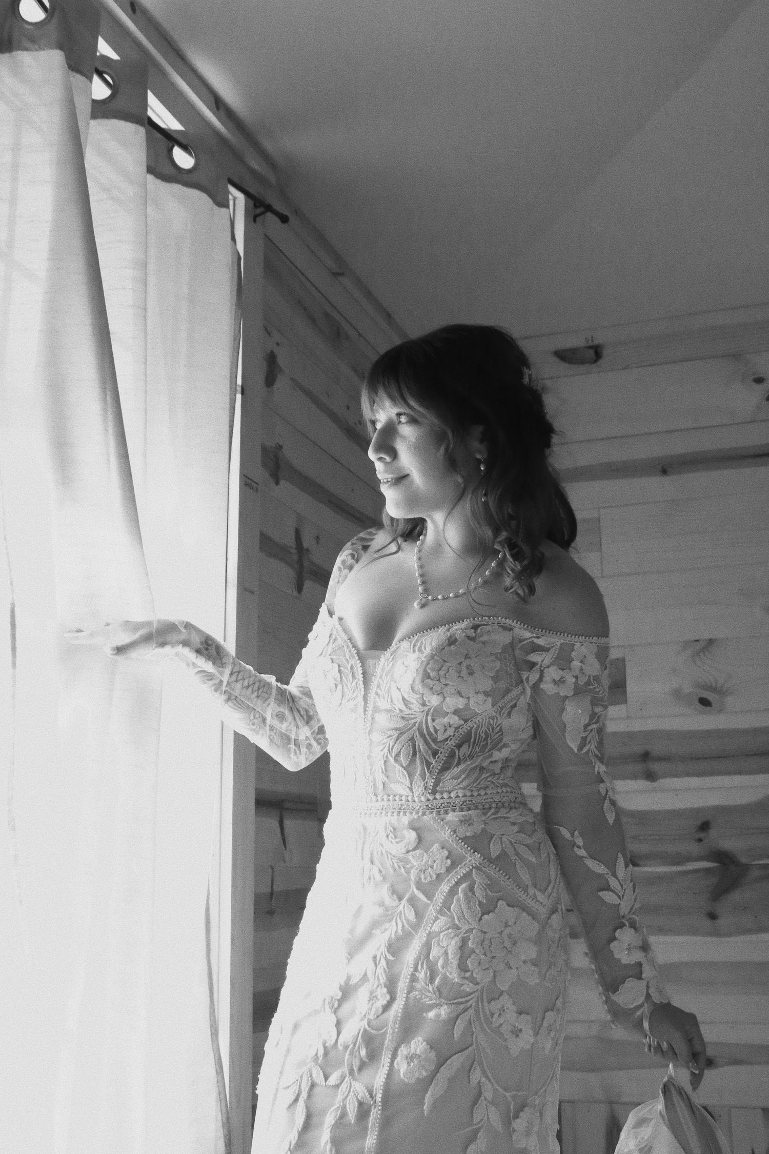 Bride in lace off-shoulder wedding gown gazes out window in black and white portrait photo.