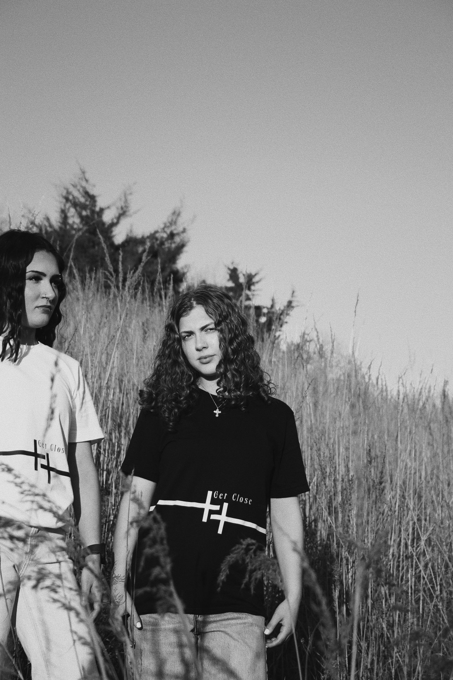 Two people in black and white photo standing in tall grass field wearing graphic tees with cross designs.
