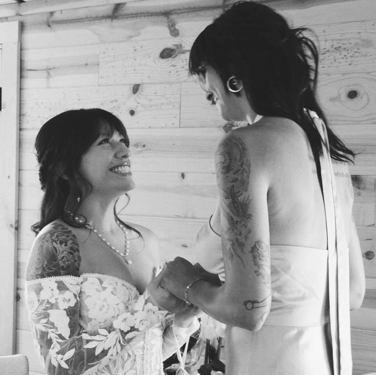 Two brides in wedding dresses hold hands and smile at each other in a black and white photo.