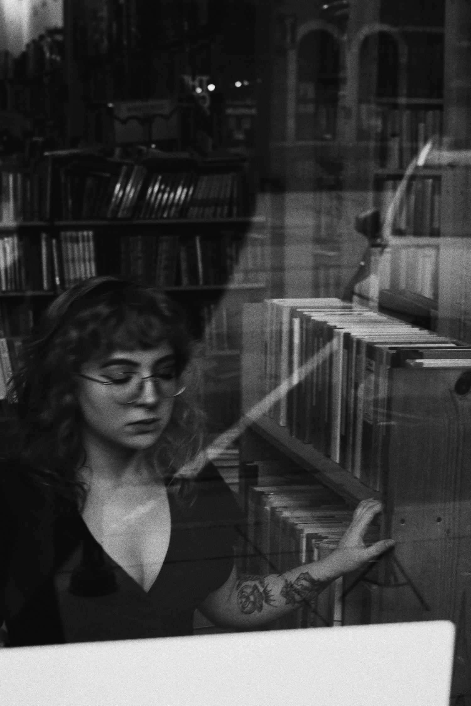 Woman with glasses and tattoos browsing books in a library, black and white photo.