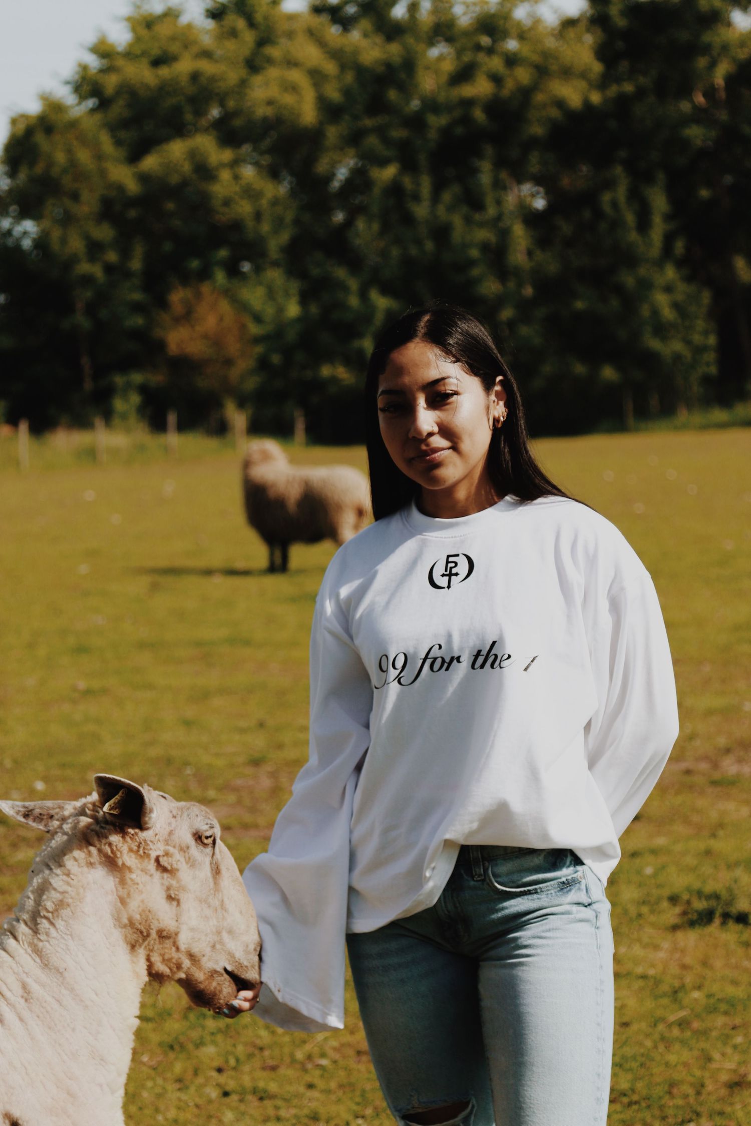 Woman in white long-sleeve shirt and jeans smiling beside a sheep in a green farm field.