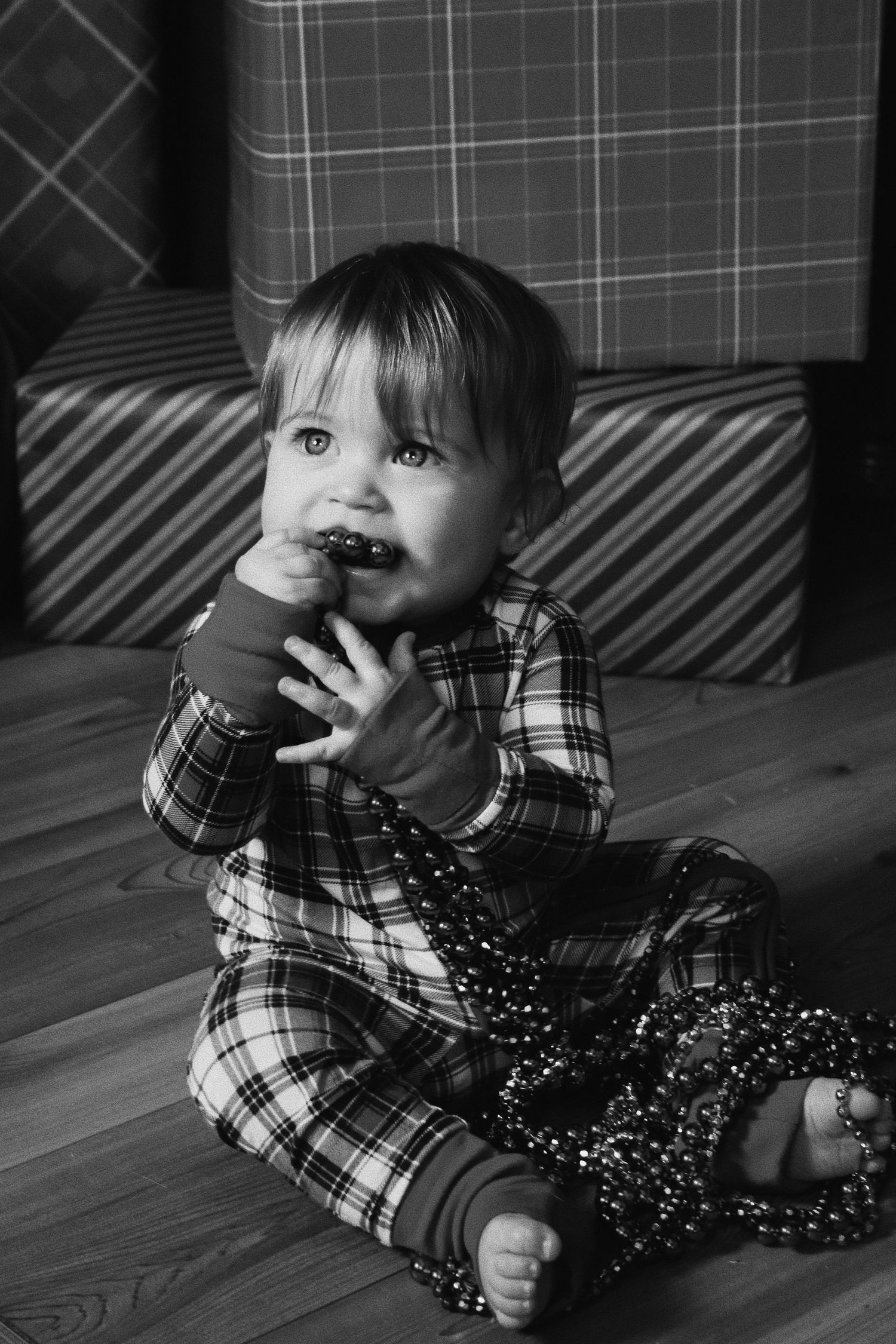 Black and white photo of a baby in plaid pajamas sitting on floor chewing beads near wrapped Christmas gifts.