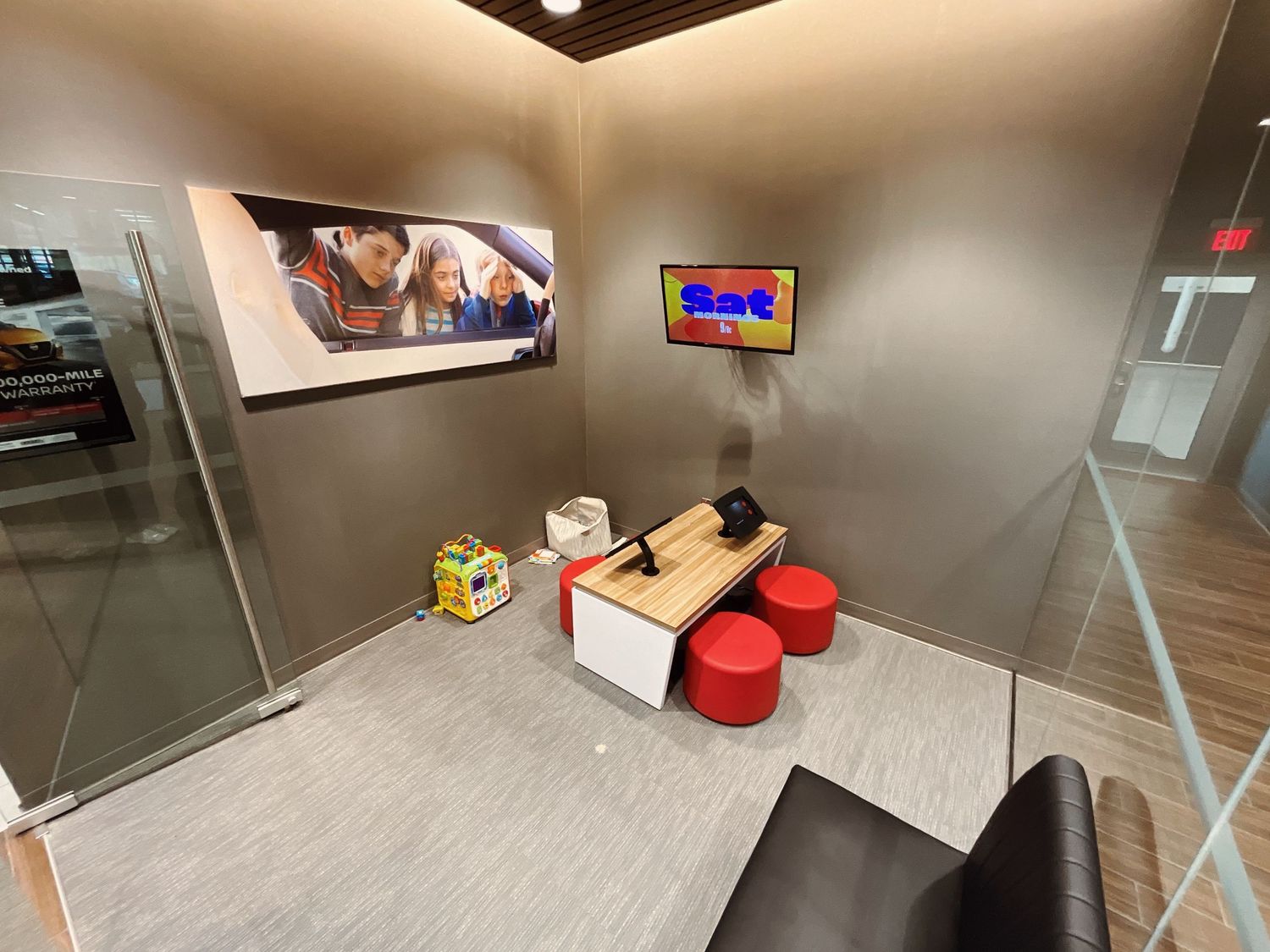Modern office kids corner with wall-mounted TV, wooden bench, red stools, and toys in a glass-enclosed play area.