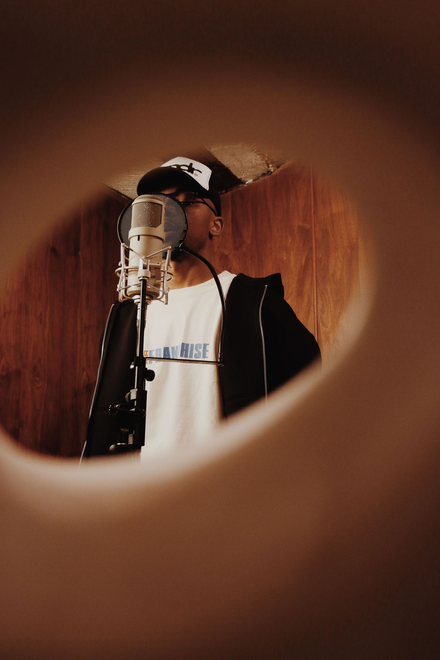 Artist recording in a professional studio booth, singing into a condenser microphone while wearing a cap and white tee.