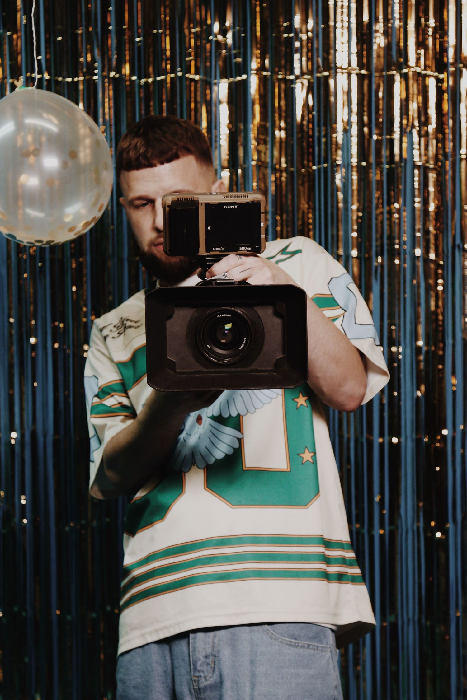 Man holding a Sony video camera at a party with gold tinsel curtain backdrop and balloons.