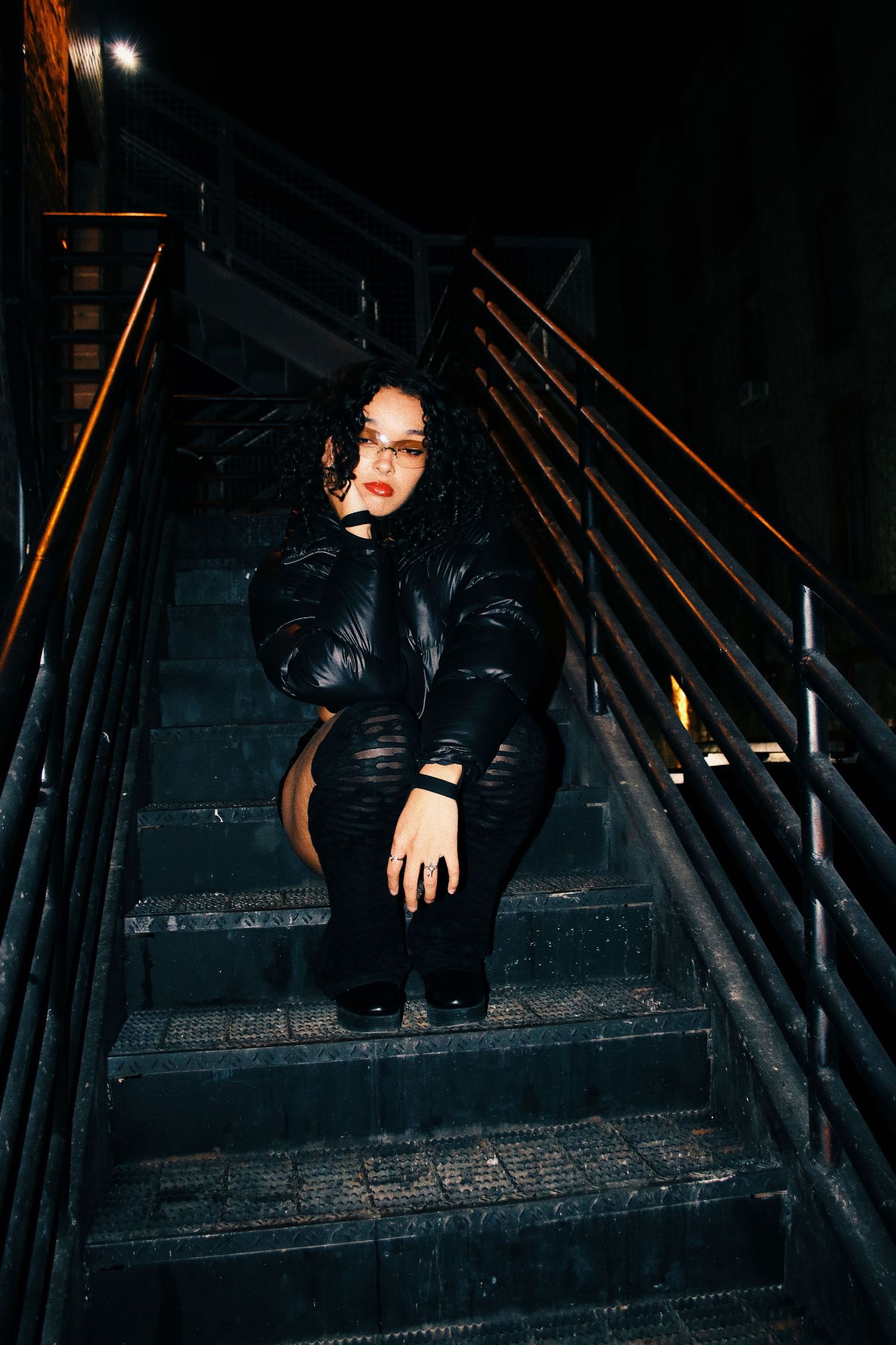 Woman in black puffer jacket sitting on dark metal stairs at night, moody urban setting.